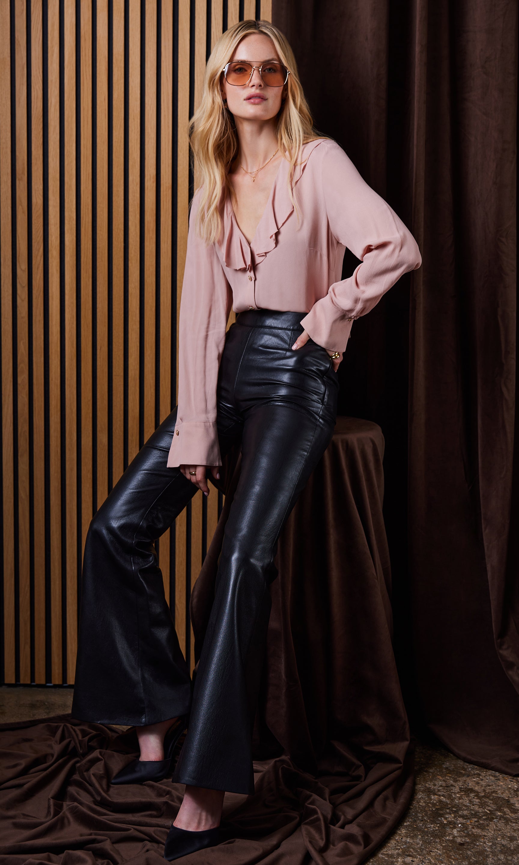 Woman wearing a pink blouse and black leather pants against a brown curtain background