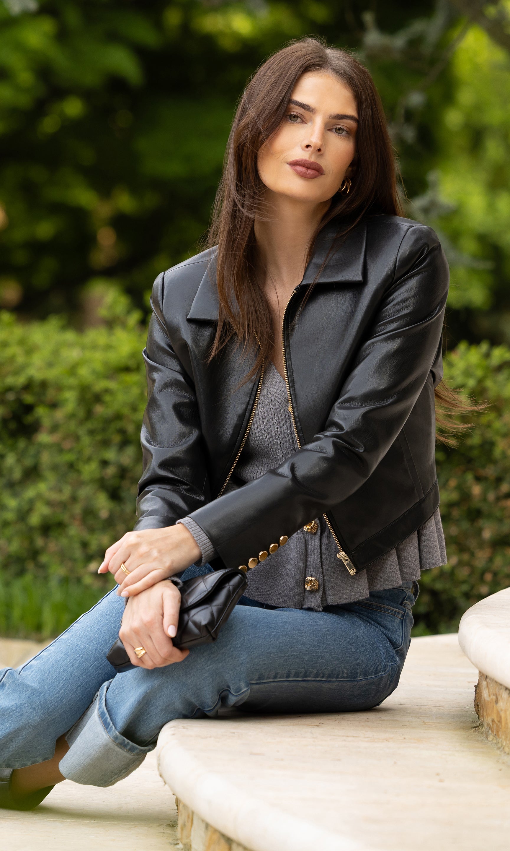 image of model sitting on a step outdoors wearing the Lola vegan leather jacket in black over a grey cardigan and blue jeans