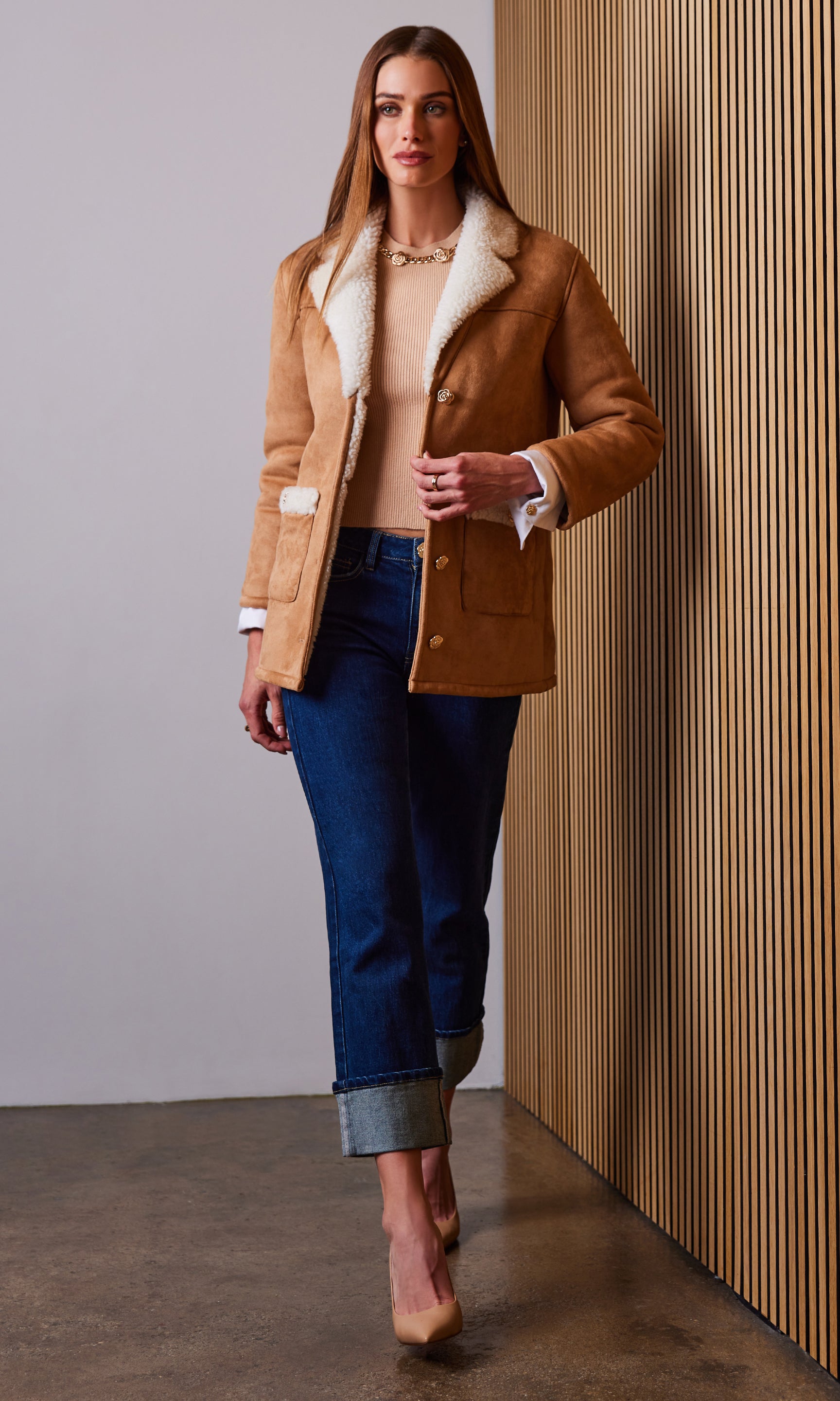 Woman wearing a tan coat with white fur trim, beige sweater, and blue jeans against a neutral wall.