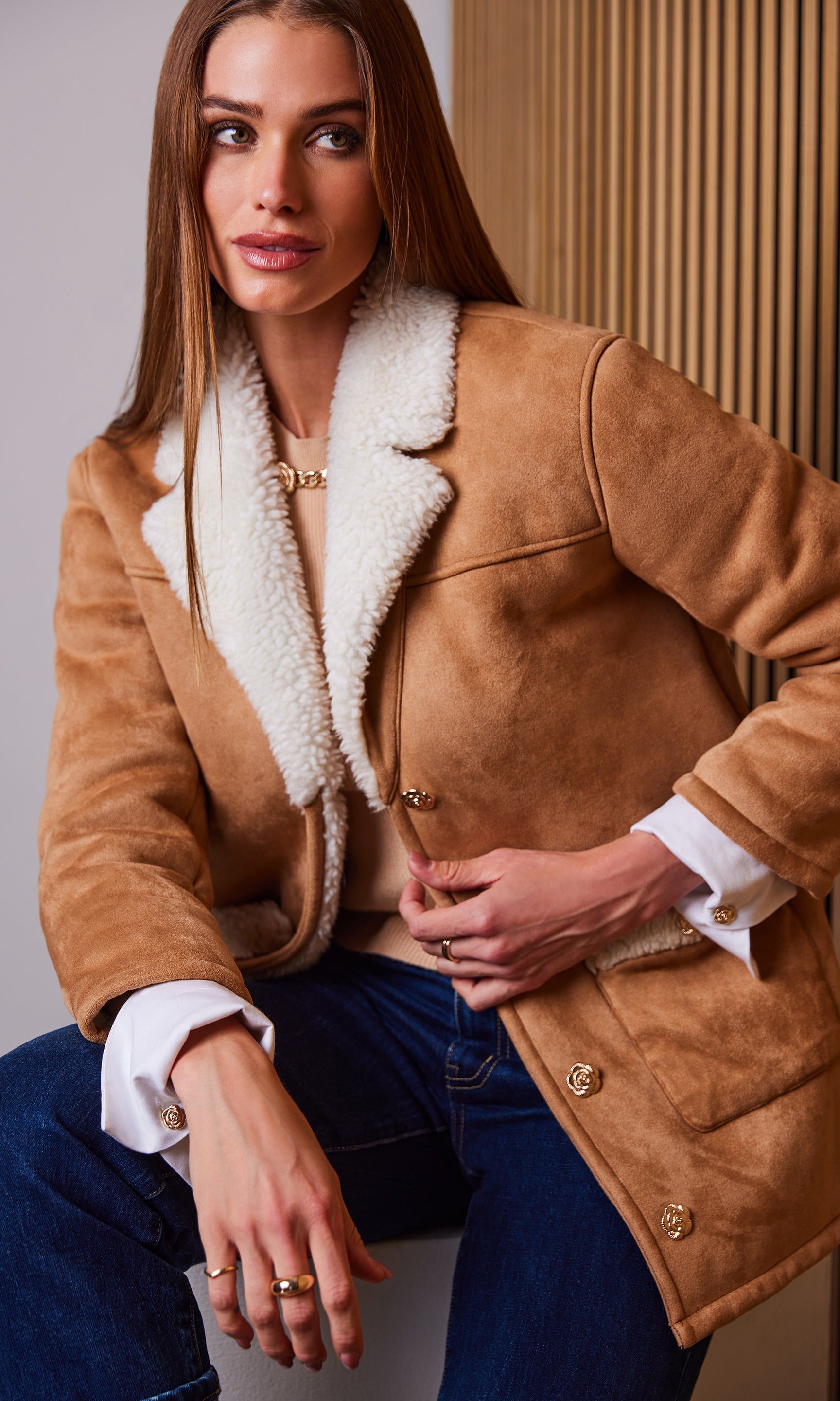 Woman wearing a brown shearling coat with white fur lining, sitting against a neutral background.