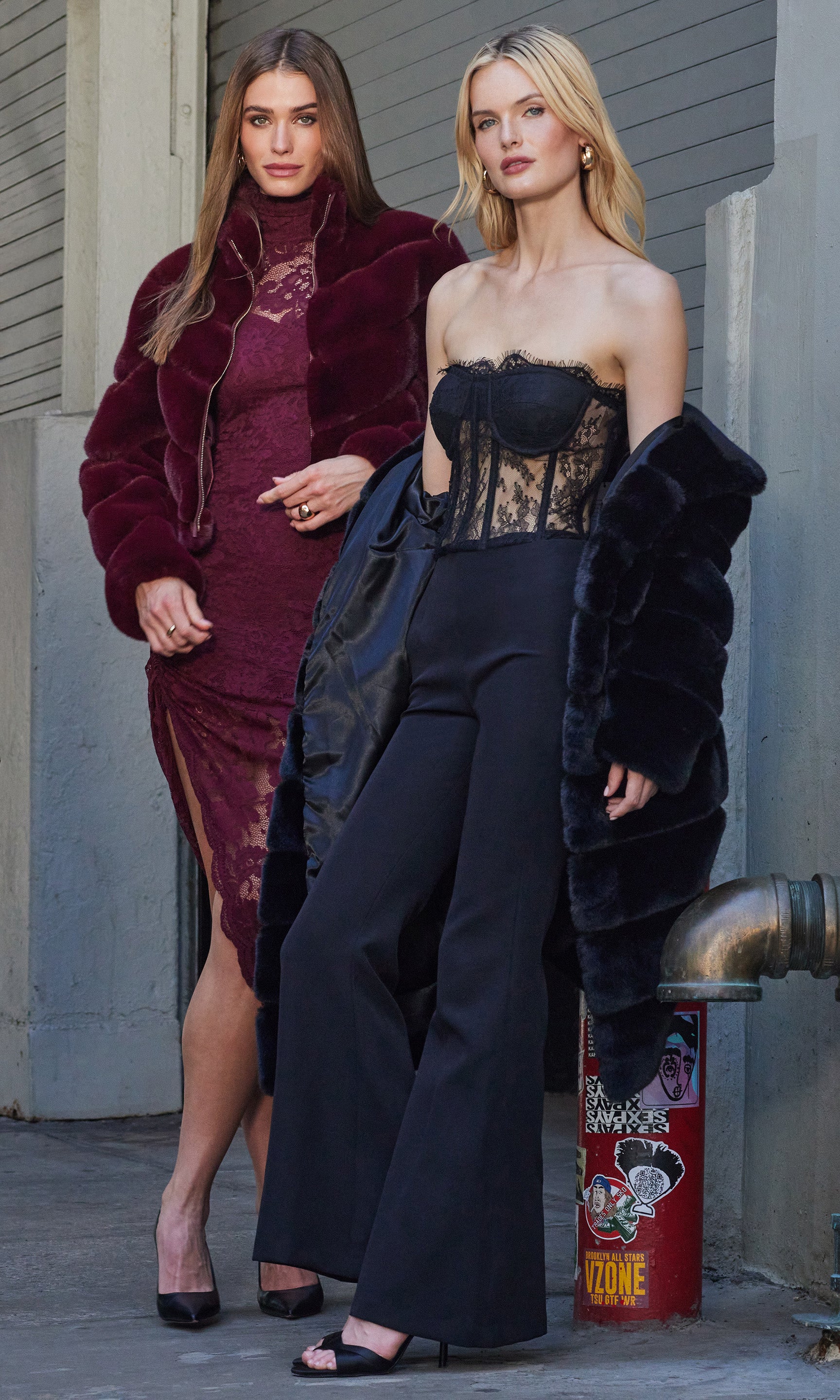 Two women in stylish outfits standing outdoors, one in a burgundy dress and the other in a black outfit with a fur coat.