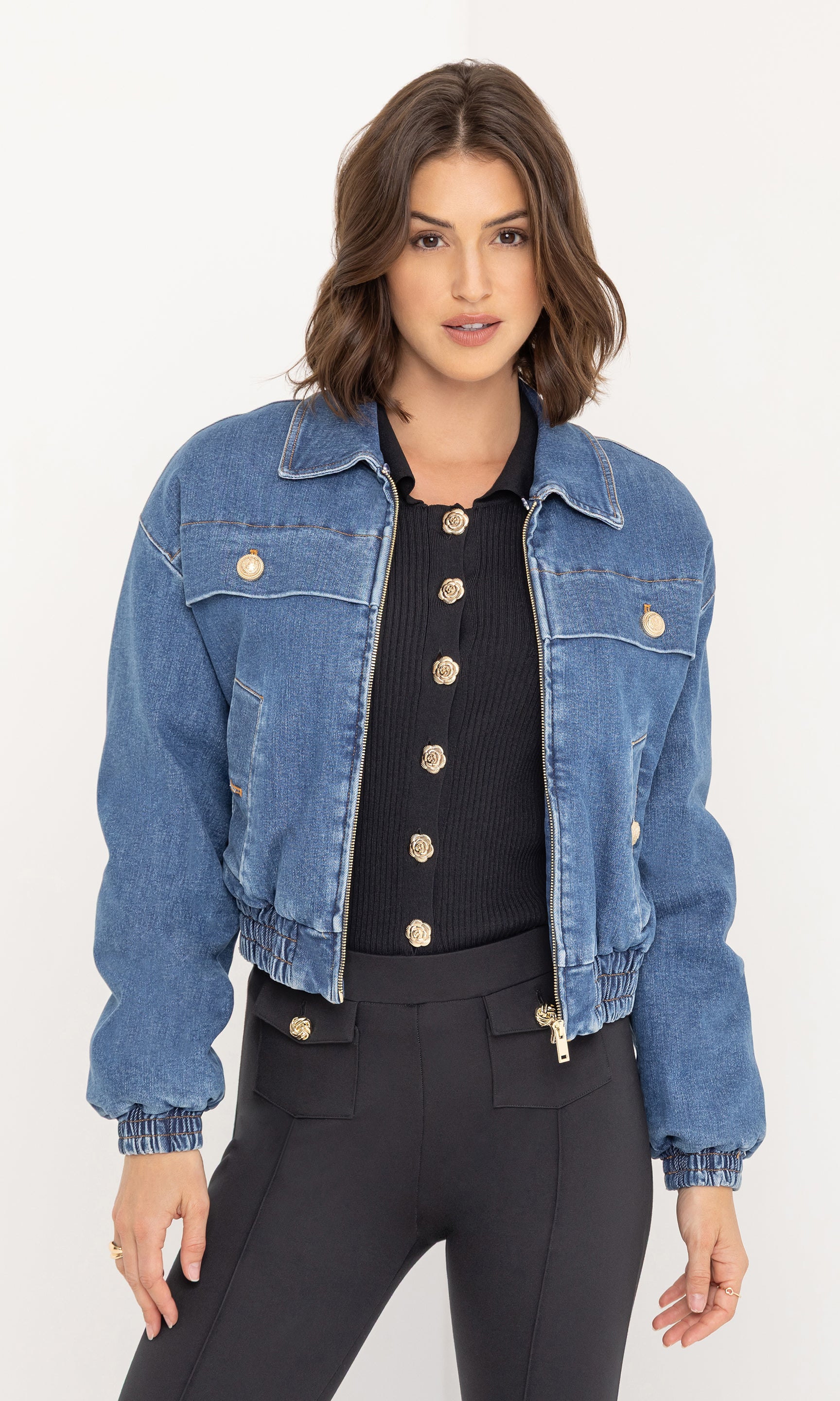 The model wears the Paco Denim Bomber without the faux fur collar, styled over a black buttoned top and black fitted trousers, in a front view showcasing the jacket’s cropped fit and gold button details.