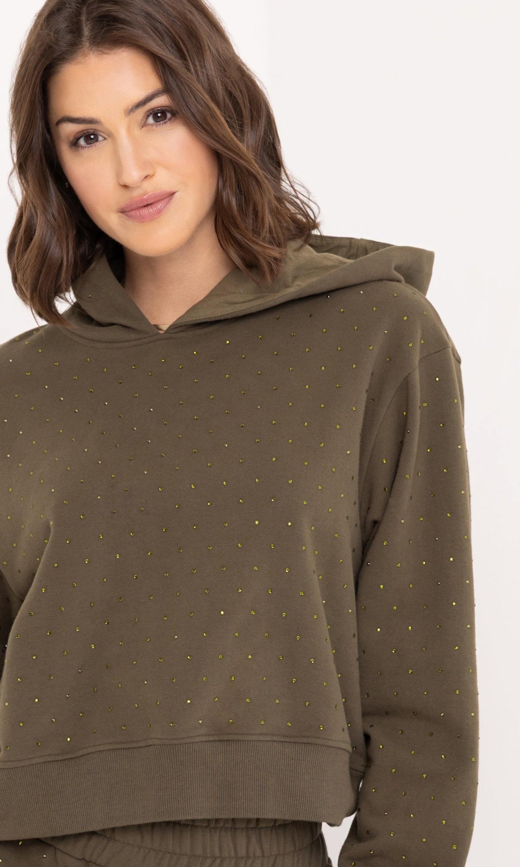 The model stands in a side view wearing the Renae Crystal Hoodie in olive, showcasing the hood and minimal crystal details.