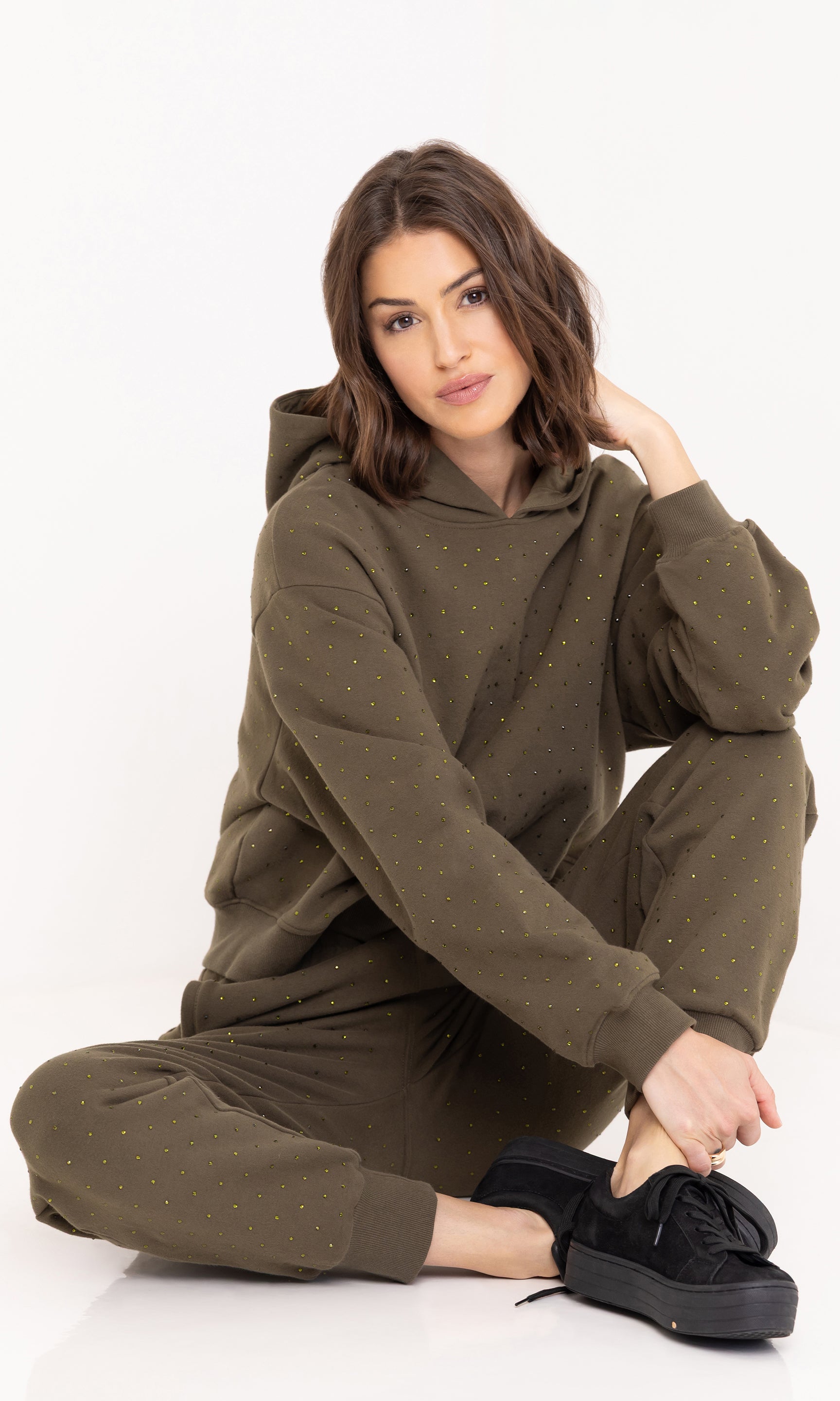 Seated pose of the model wearing the Ruel Crystal Sweatpants in olive, paired with a matching sweatshirt and sneakers.