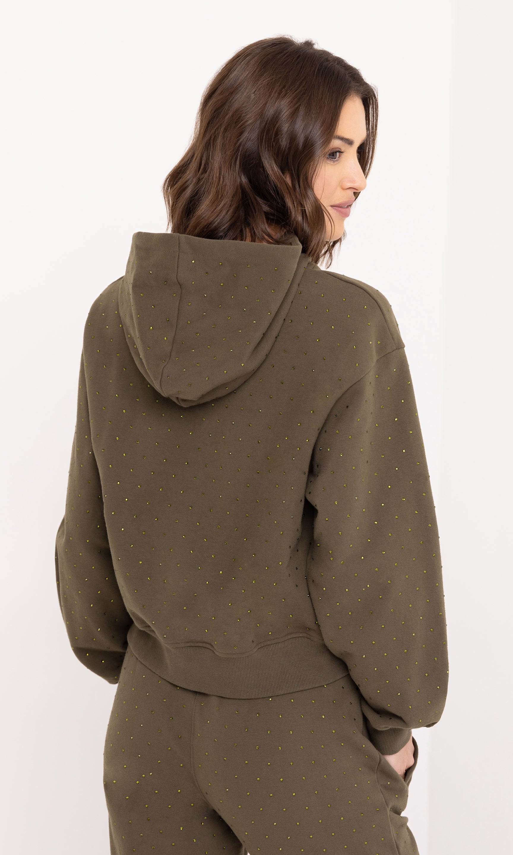 The model poses in a three-quarter view wearing the Renae Crystal Hoodie in olive, highlighting the hood and comfortable structure.