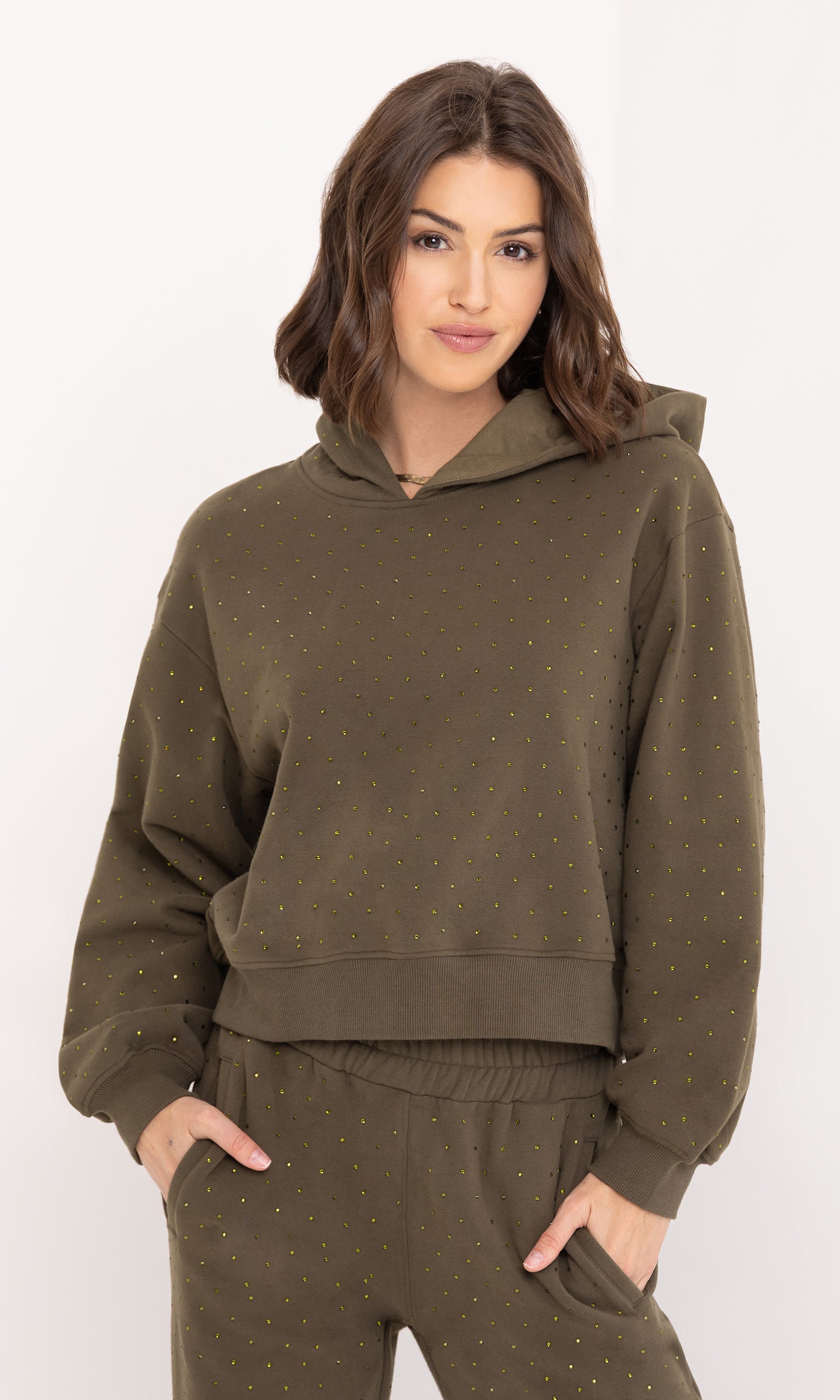 The model stands in a front view wearing the Renae Crystal Hoodie in olive, hands in pockets, emphasizing its cozy silhouette and crystal accents.