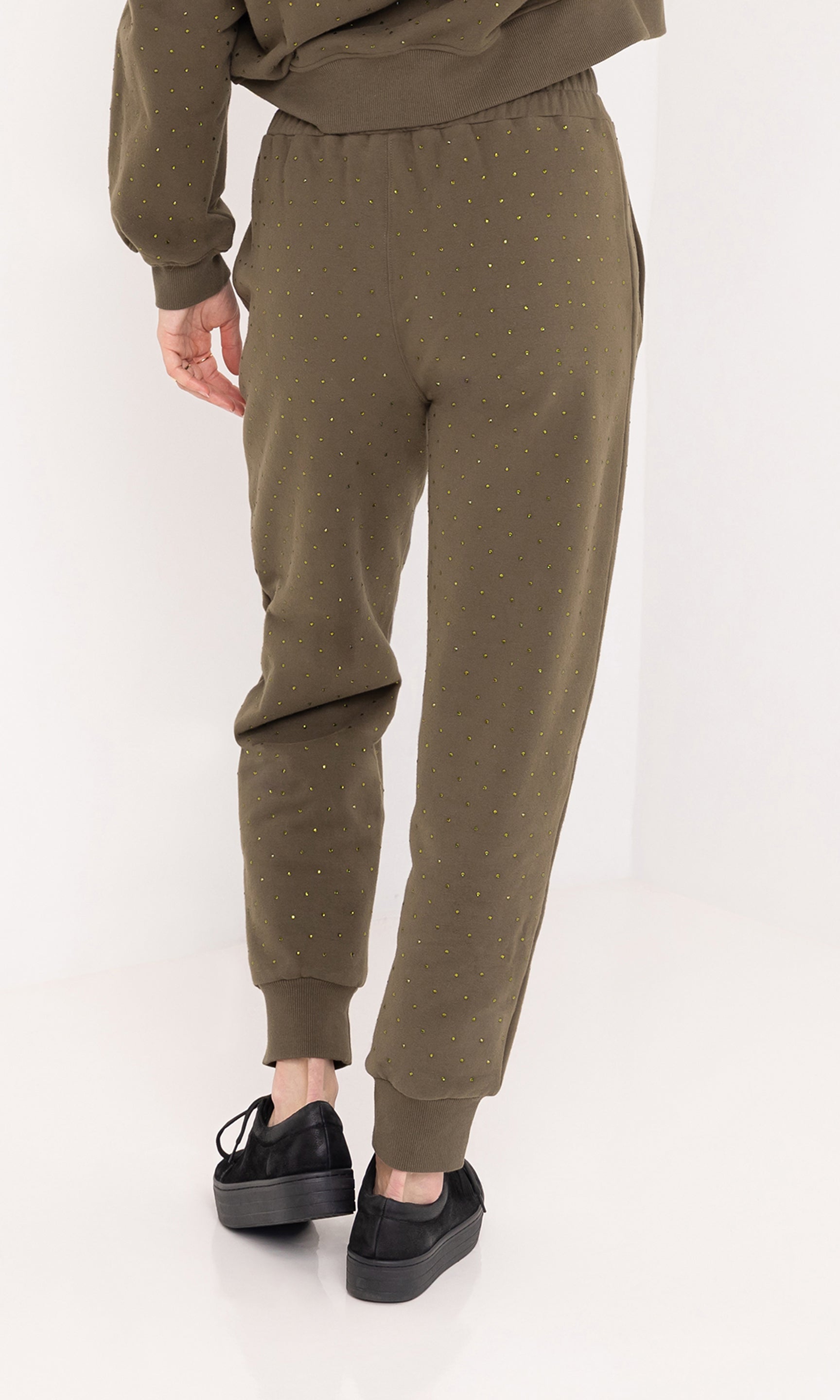 The model is shown from the rear view wearing the ruel crystal sweatpants in olive green, highlighting the slim jogger fit, elastic waistband, and gold crystal embellishments throughout.