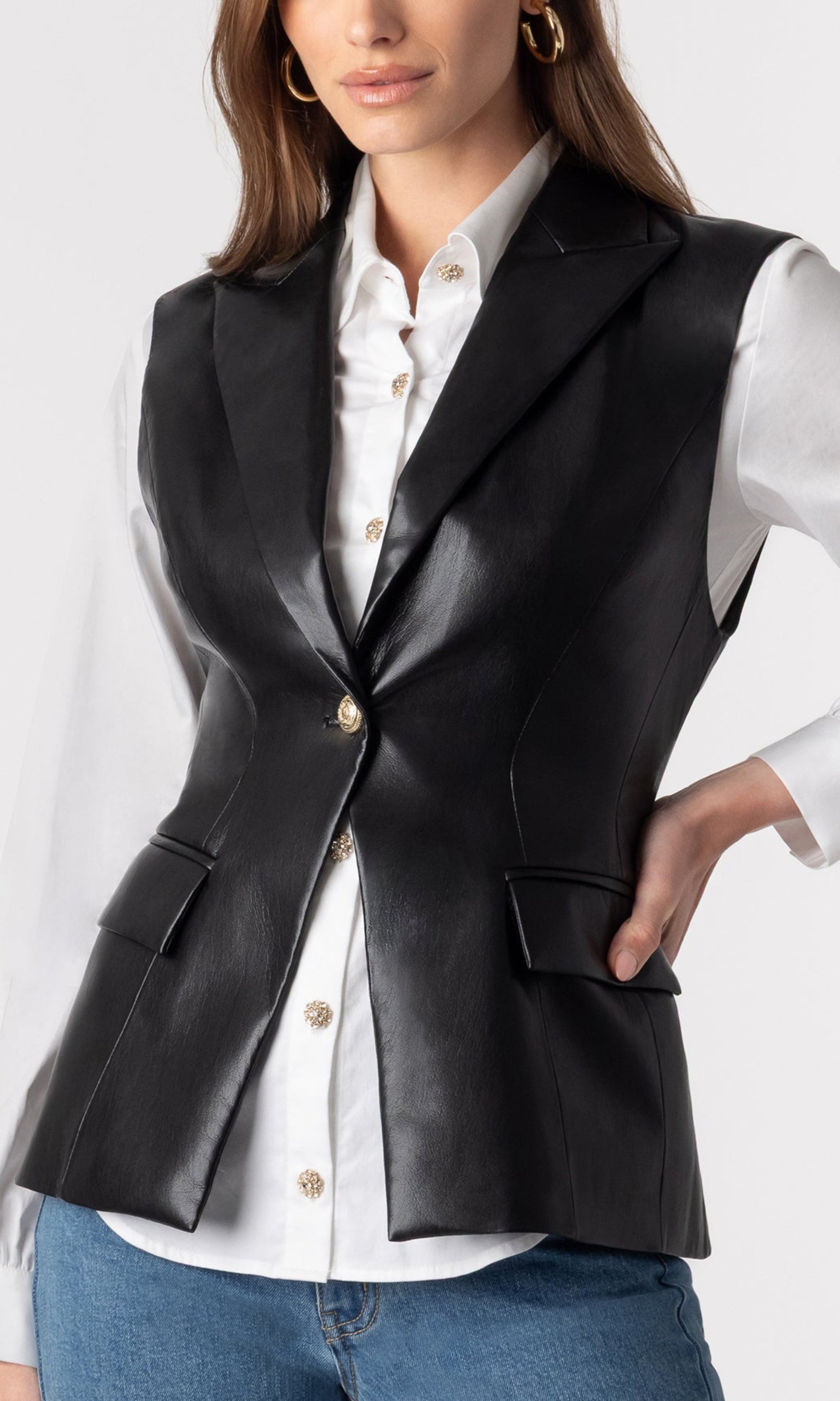 zoomed in image of model wearing the vegan leather Rialto sleeveless blazer over a white button-down blouse and paired with blue jeans, showcasing the structure of the blazer with a singular front button closure