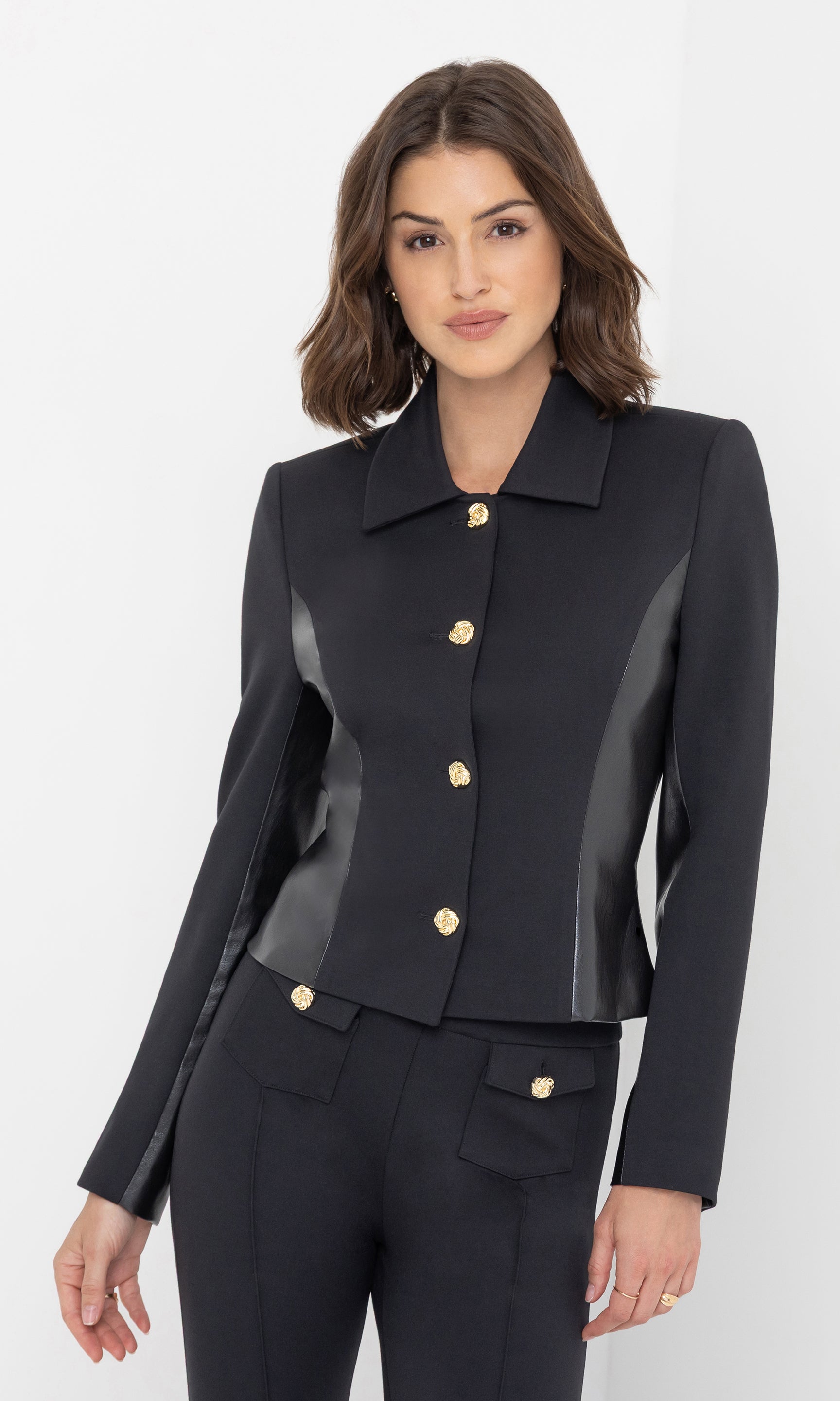 The model wears the Robby Vegan Leather Combo Ponte Jacket in black, shown in a front view with one hand resting by her side, highlighting the structured silhouette, vegan leather side panels, and gold button detailing.