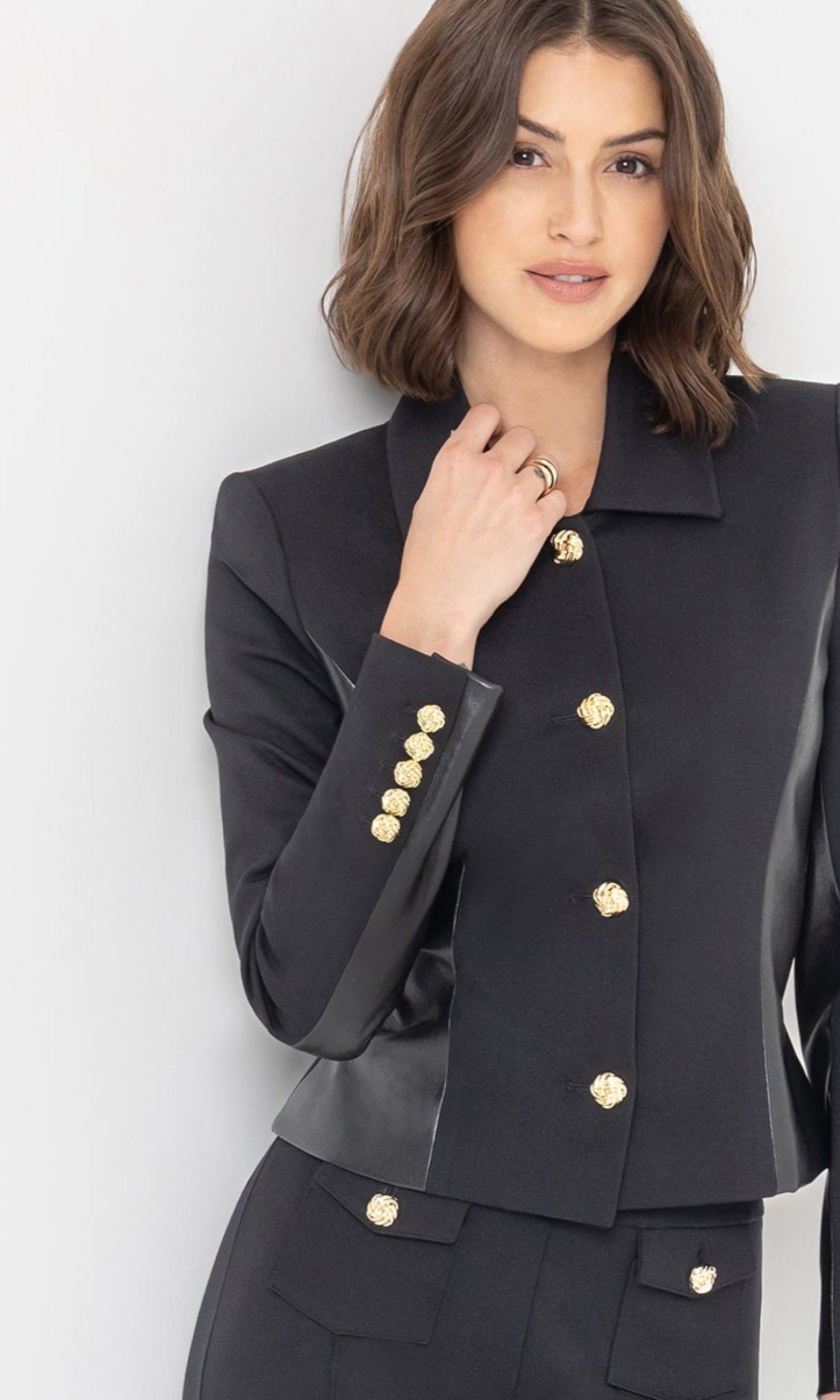 The model showcases the Robby Vegan Leather Combo Ponte Jacket in black from a three-quarter front view, emphasizing the tailored fit and gold button accents.