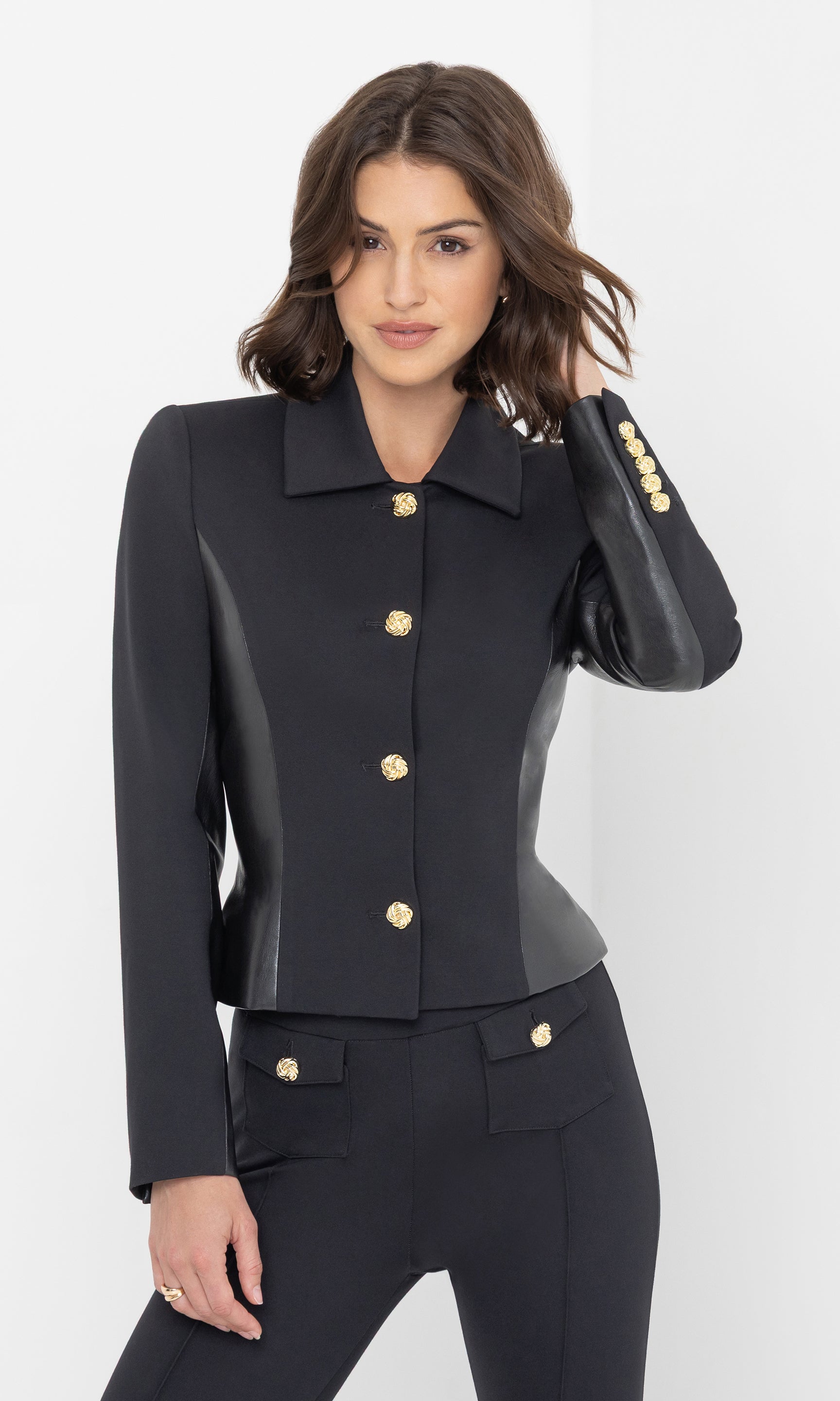 The model wears the Robby Vegan Leather Combo Ponte Jacket in black, posing in a front view with hands in pockets, highlighting the gold button detailing.