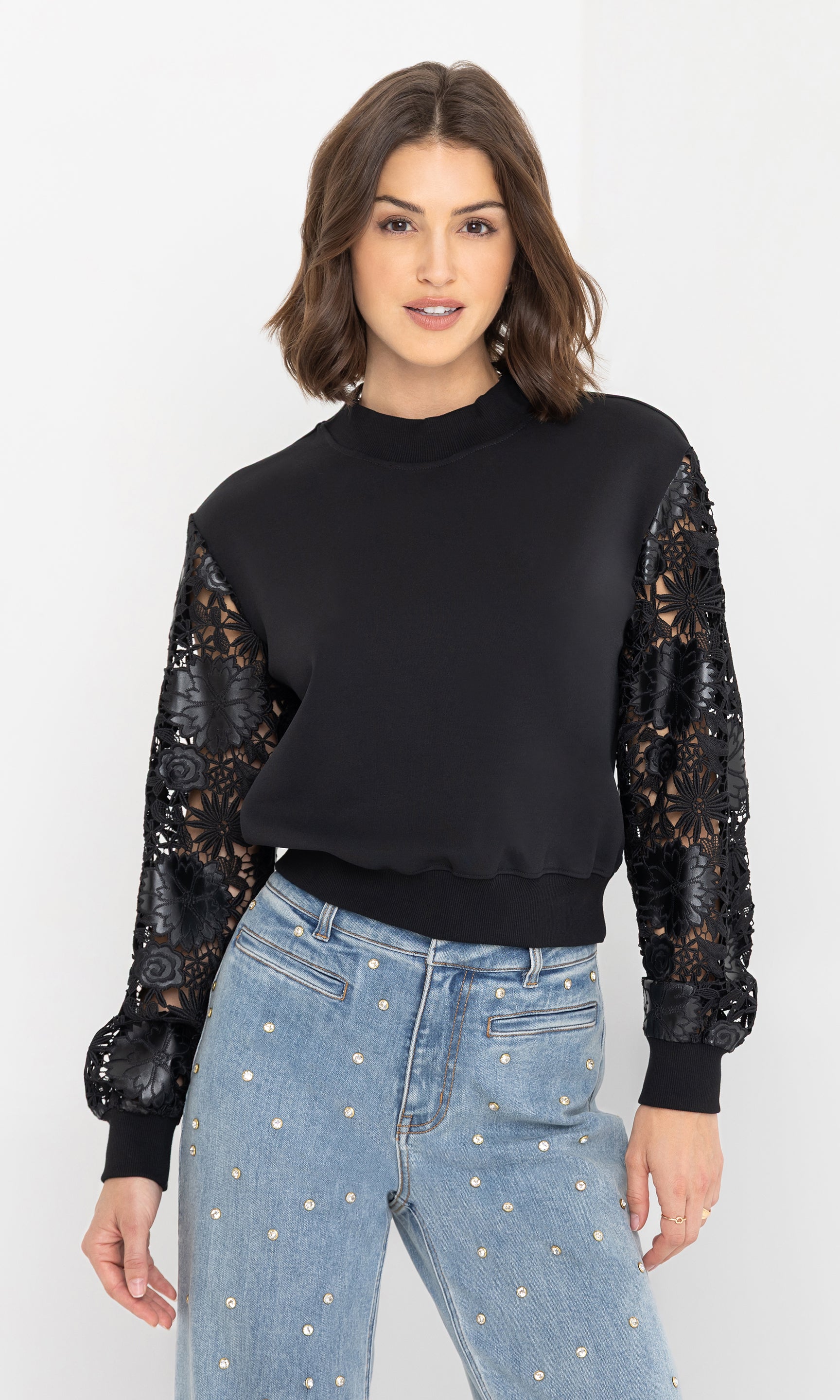 The model poses in a front view wearing the theron guipure combo sleeve sweatshirt in black, styled with light-wash crystal-embellished jeans.