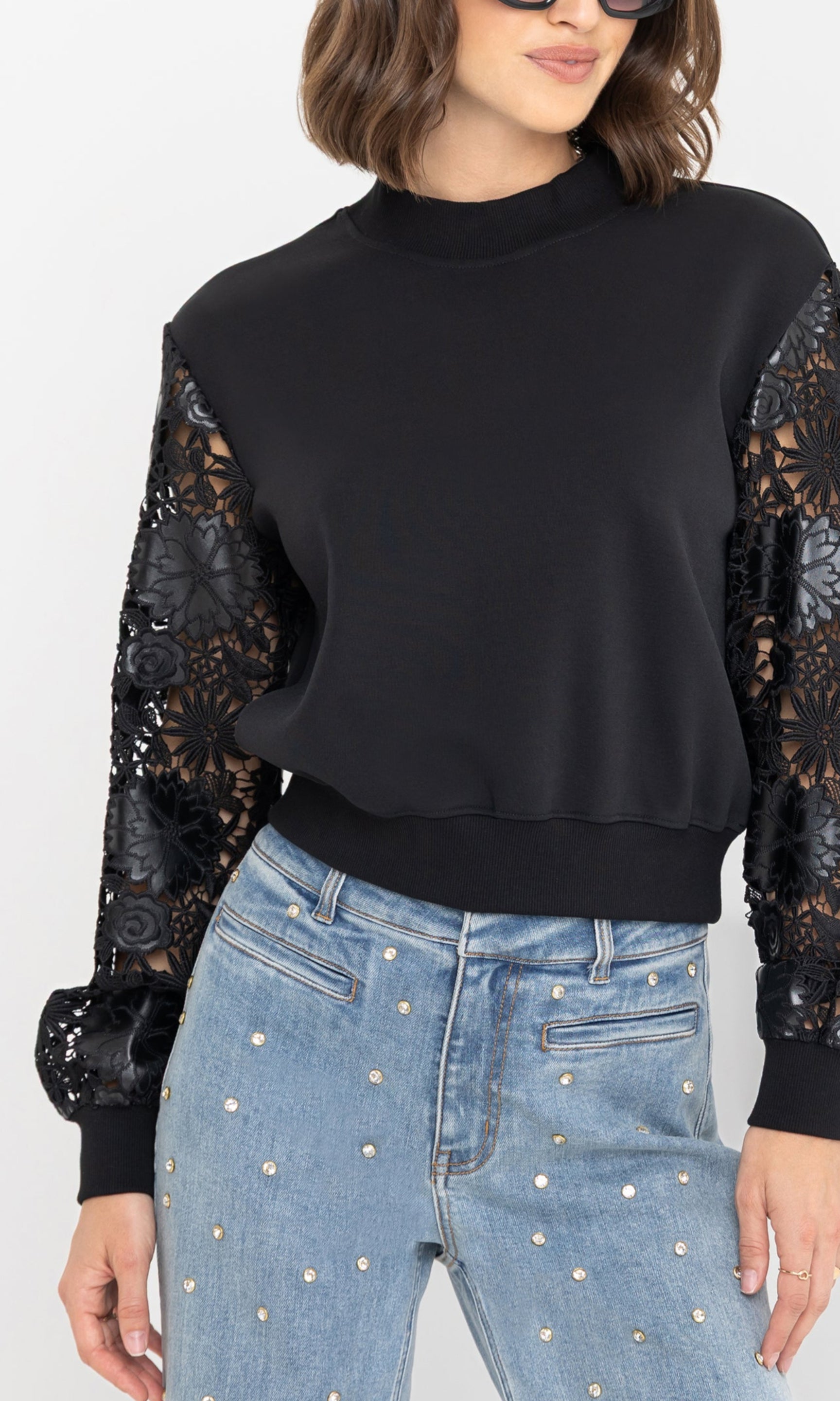 Close-up front view of the Theron Guipure Combo Sleeve Sweatshirt in black, highlighting the sheer lace sleeves and paired with light-wash crystal-embellished jeans.
