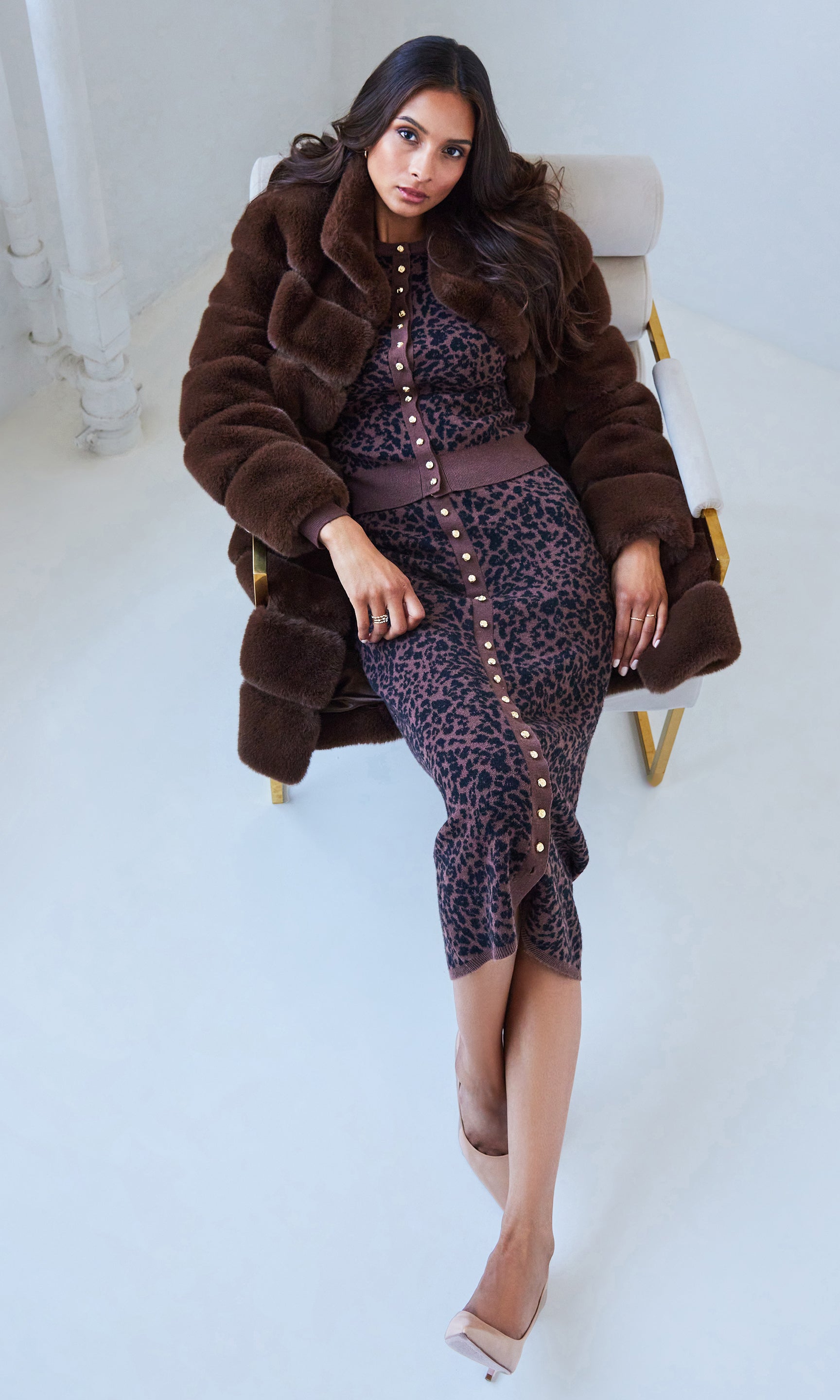 Woman wearing a brown fur coat and animal patterned outfit sitting on a chair.