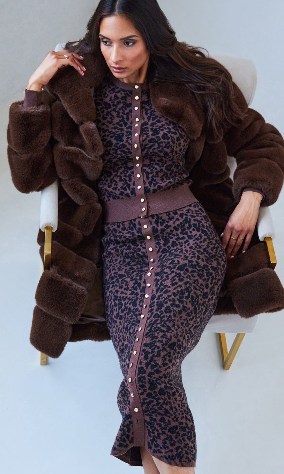 Woman wearing a brown fur coat over a leopard print top and skirt in a minimalistic setting.