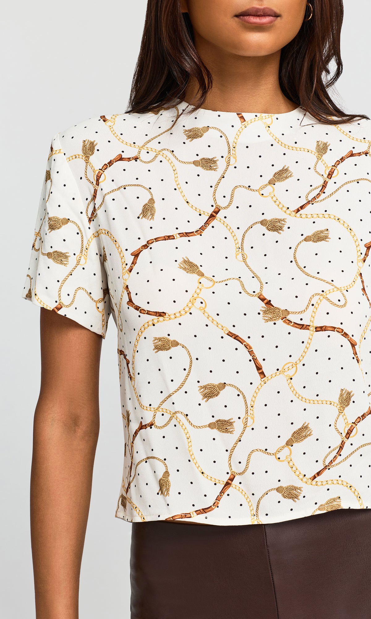 Close-up of a short-sleeve crop top with an off-white base, black polka dots, and a gold and brown chain-link and tassel pattern.