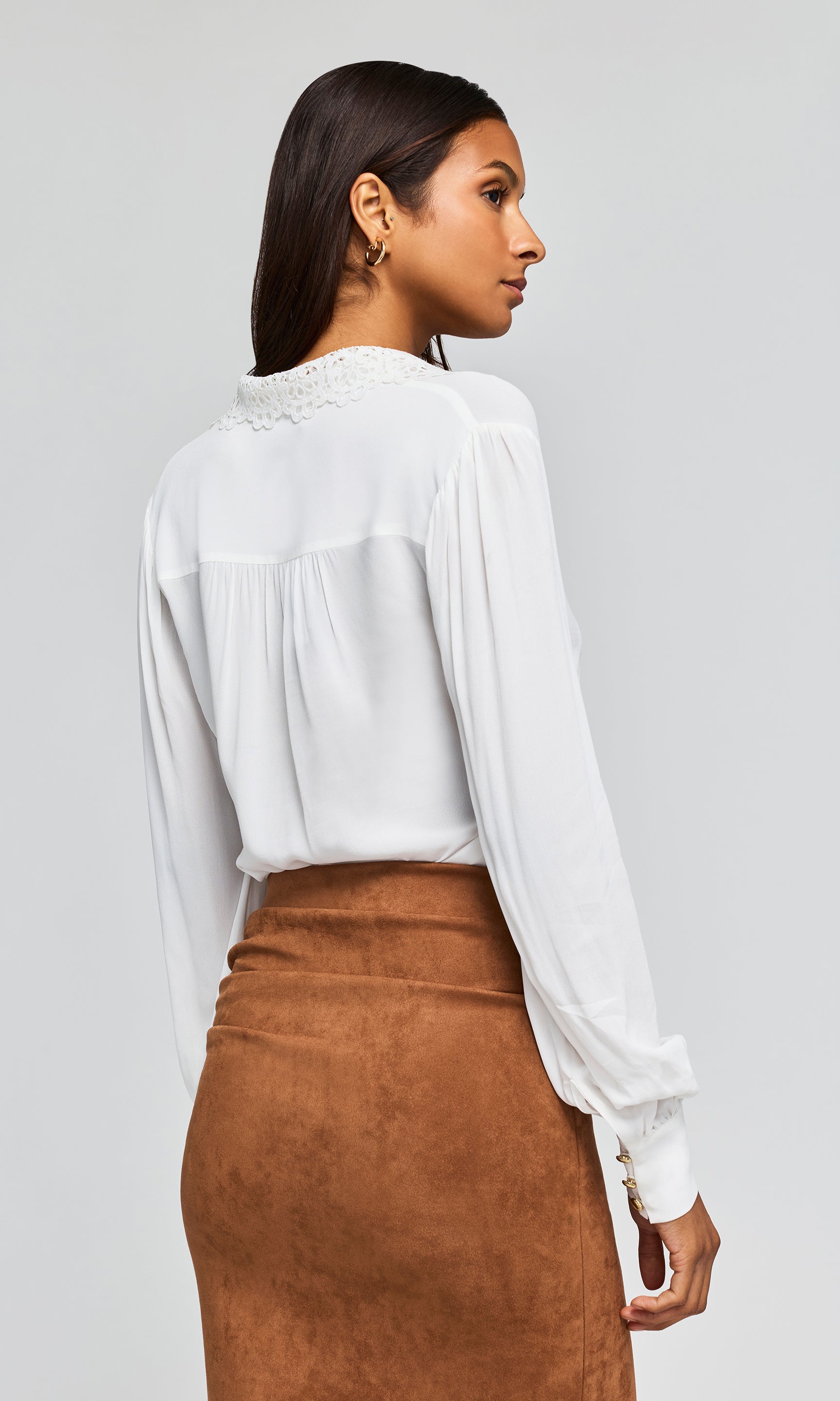 Back view of model wearing a white blouse with  decorative lace detail at the collar, paired with a ruched brown skirt.