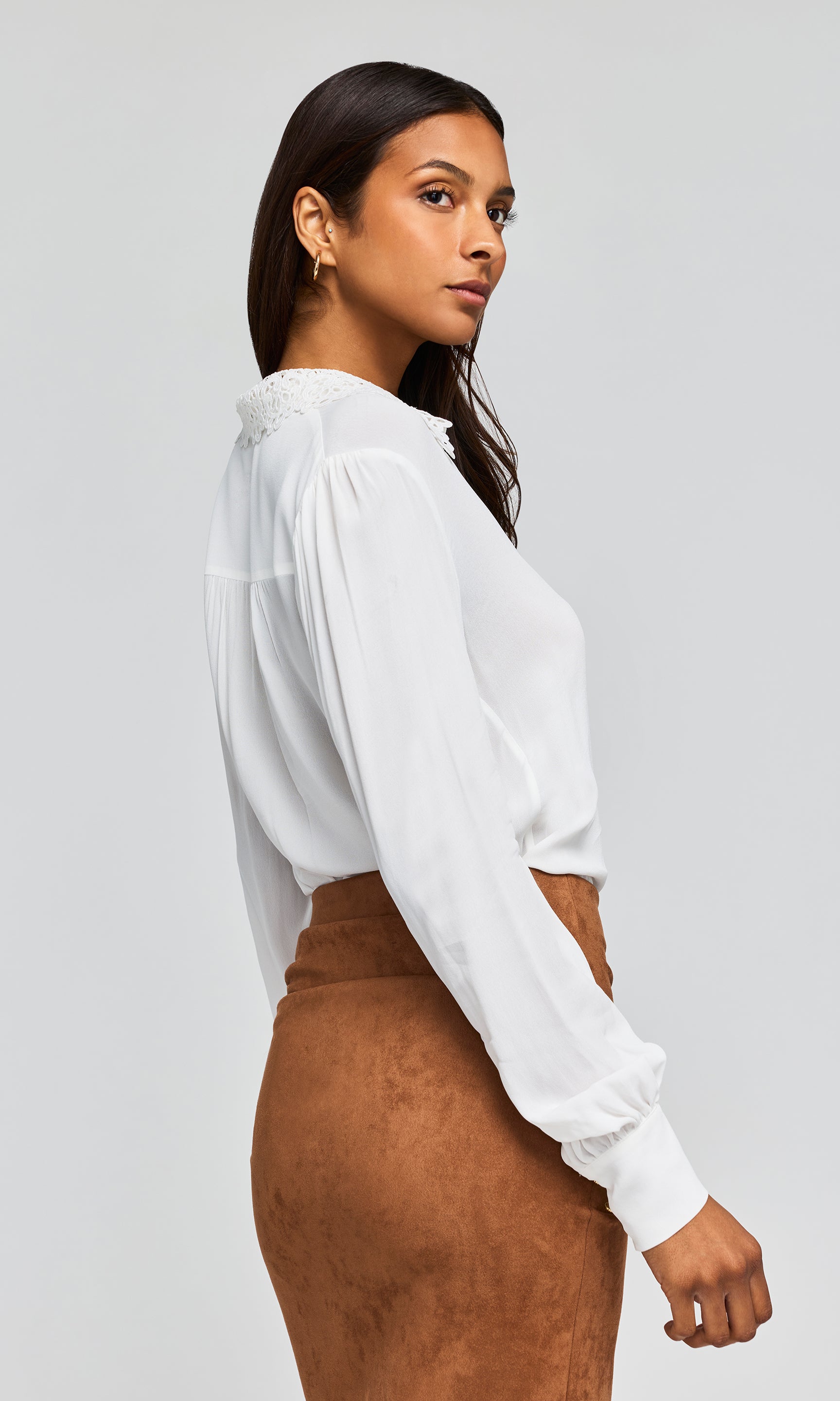 Side view of model wearing a white blouse with  decorative lace detail at the collar, paired with a ruched brown skirt.