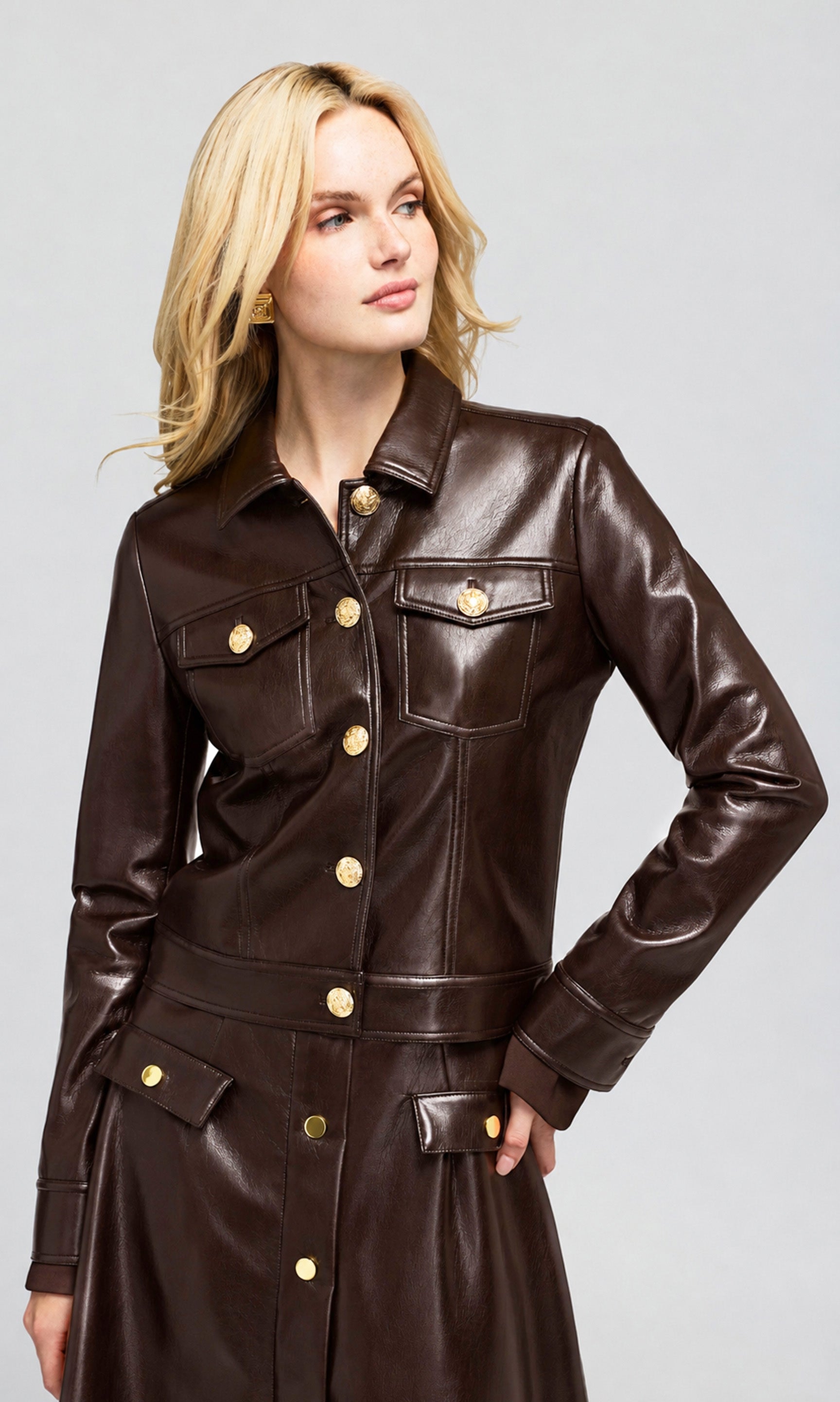A blonde model wearing a dark brown faux-leather jacket with gold-tone buttons, on a plain background.