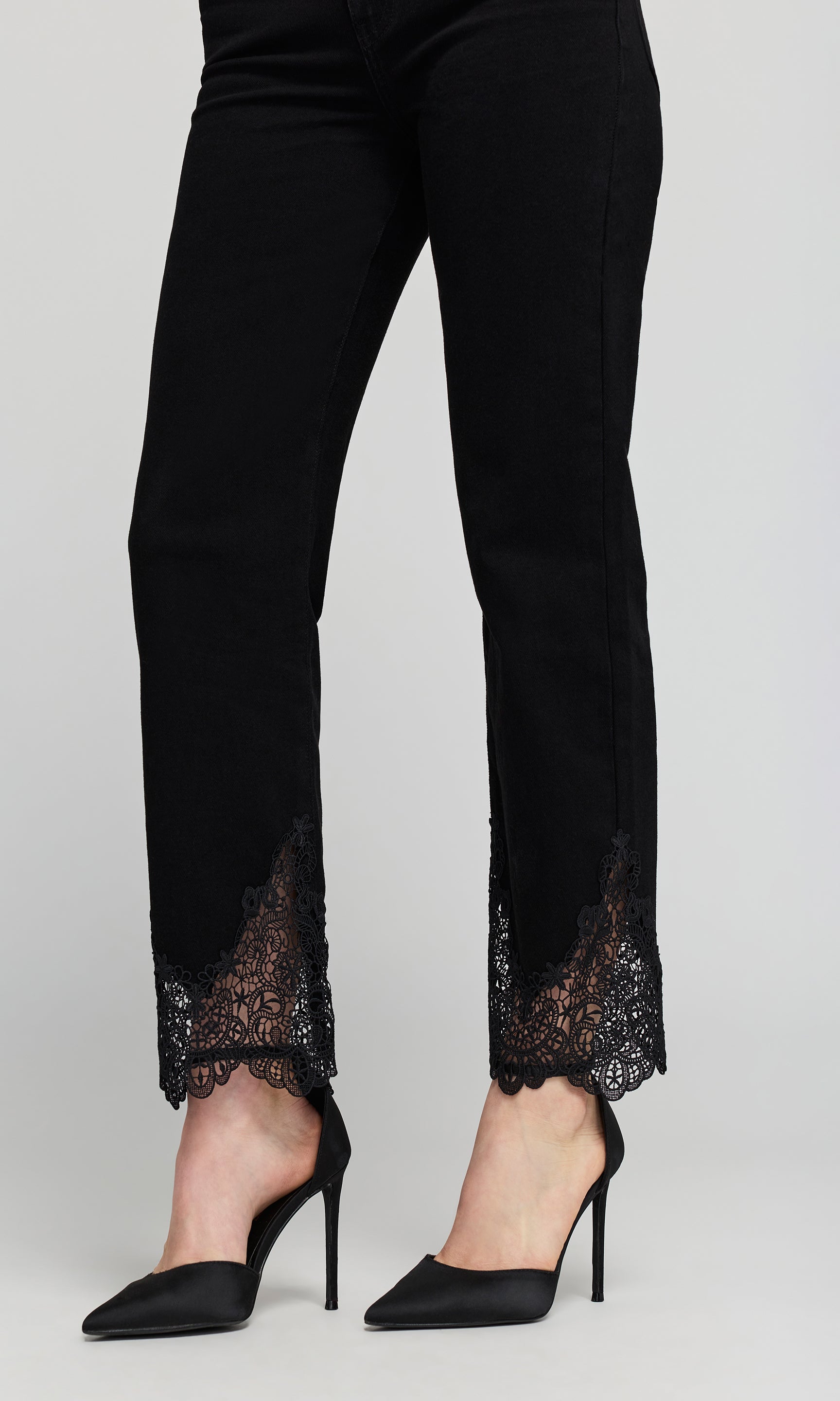 Close up of black ankle-length pants with sheer black lace trim at the hem, on a plain background.