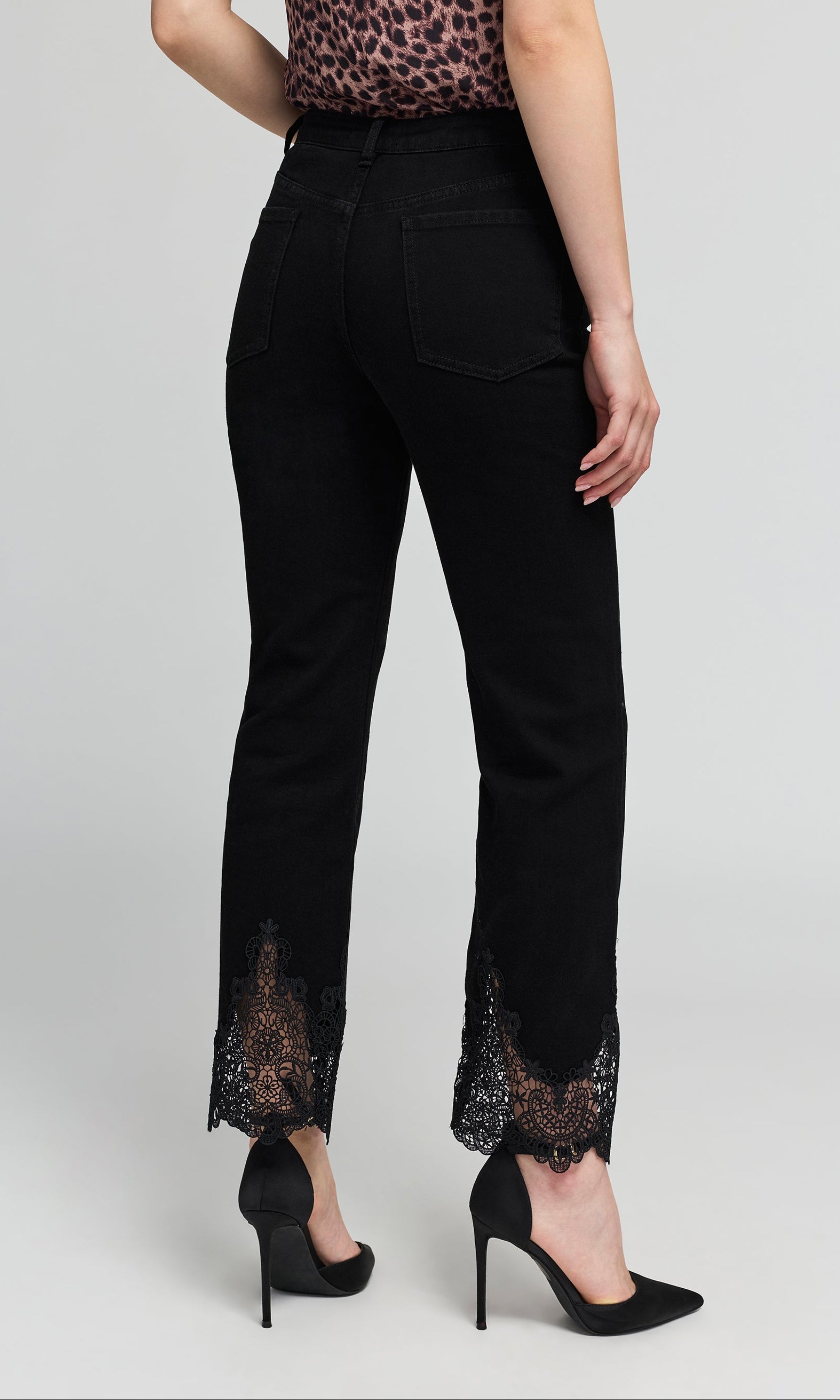 Back view of black cropped  jeans with black lace trim at the hem, shown on a model on a plain background.