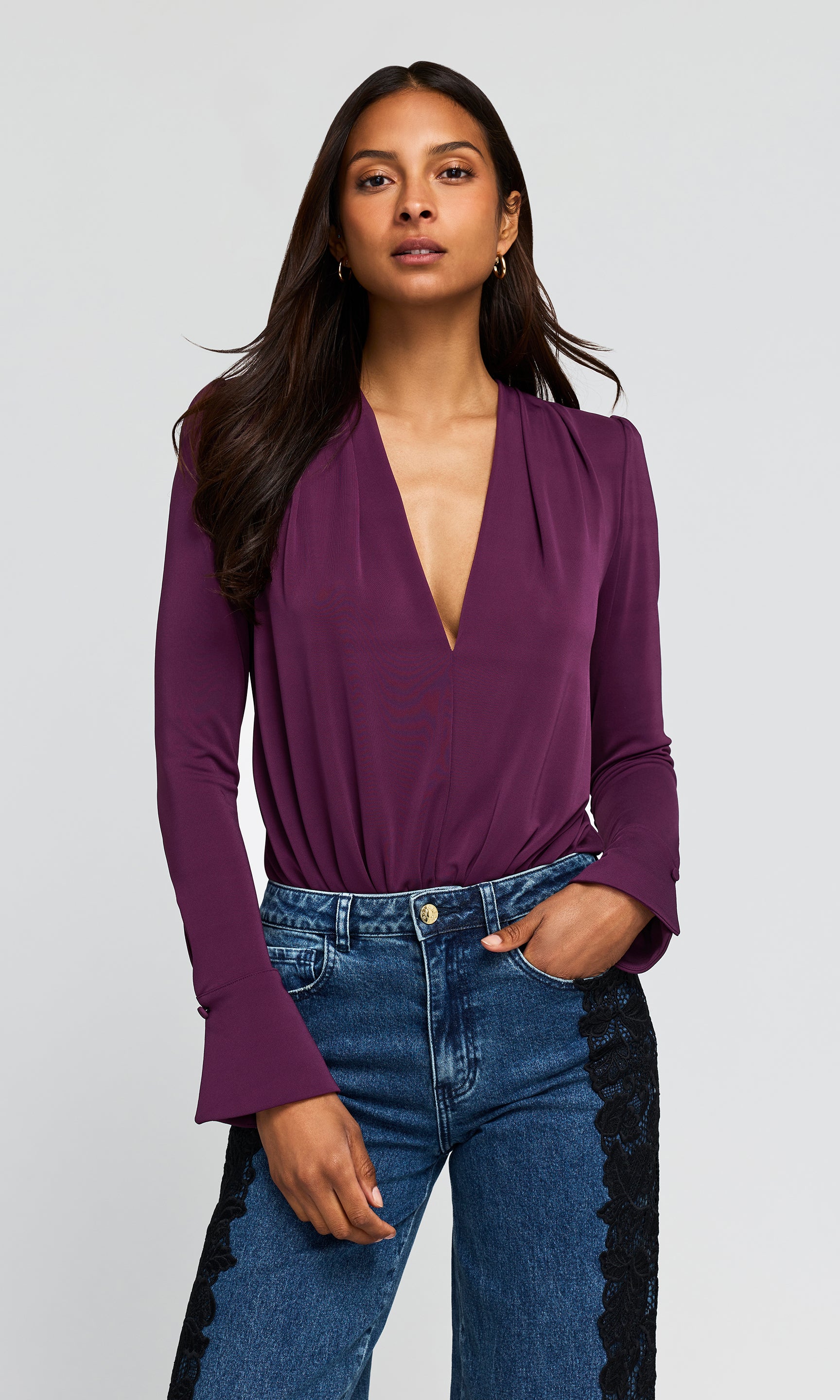 Model wearing a deep purple blouse with a plunging V-neckline and flared cuffs, paired with blue jeans.