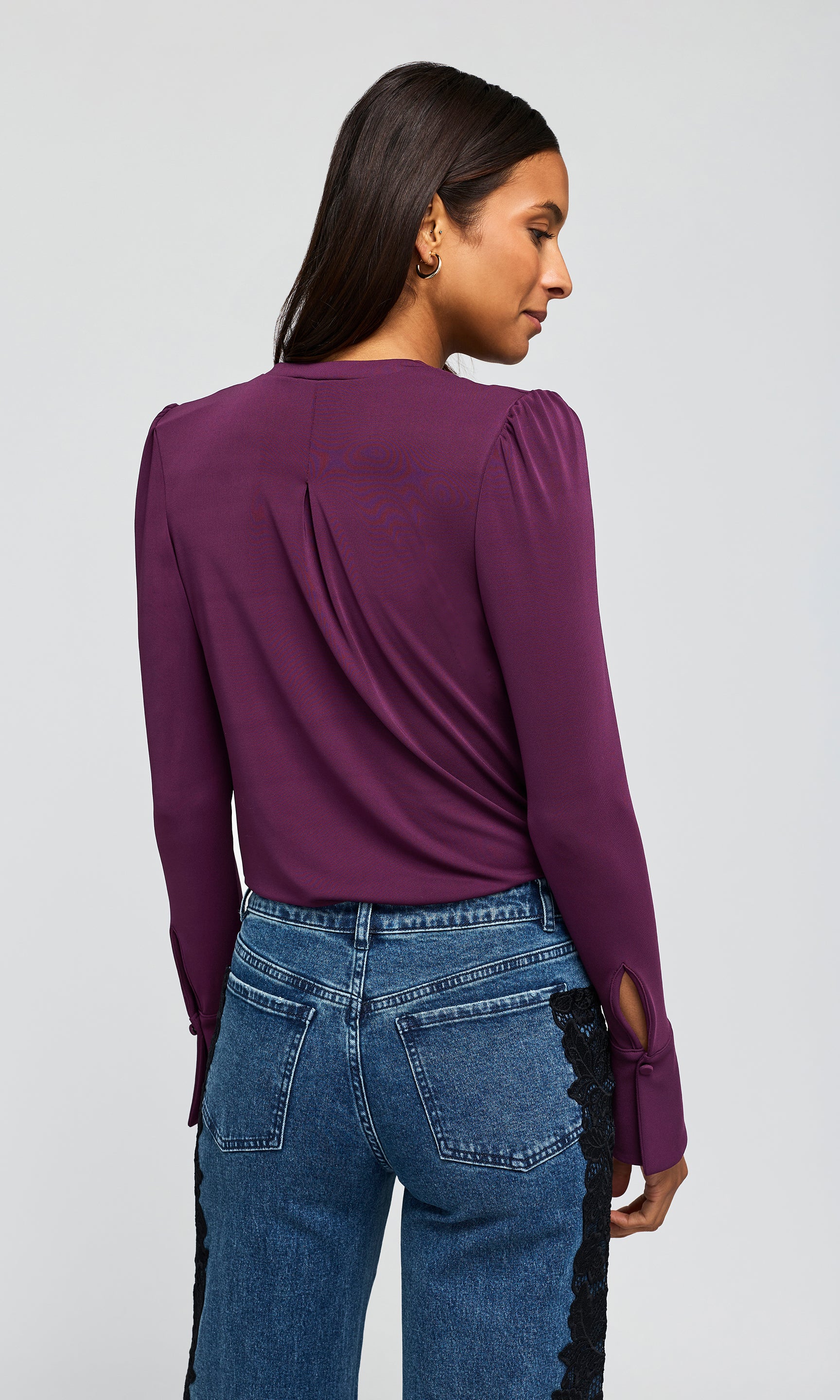 Back view of mdel wearing a deep purple blouse with flared cuffs, paired with blue jeans.
