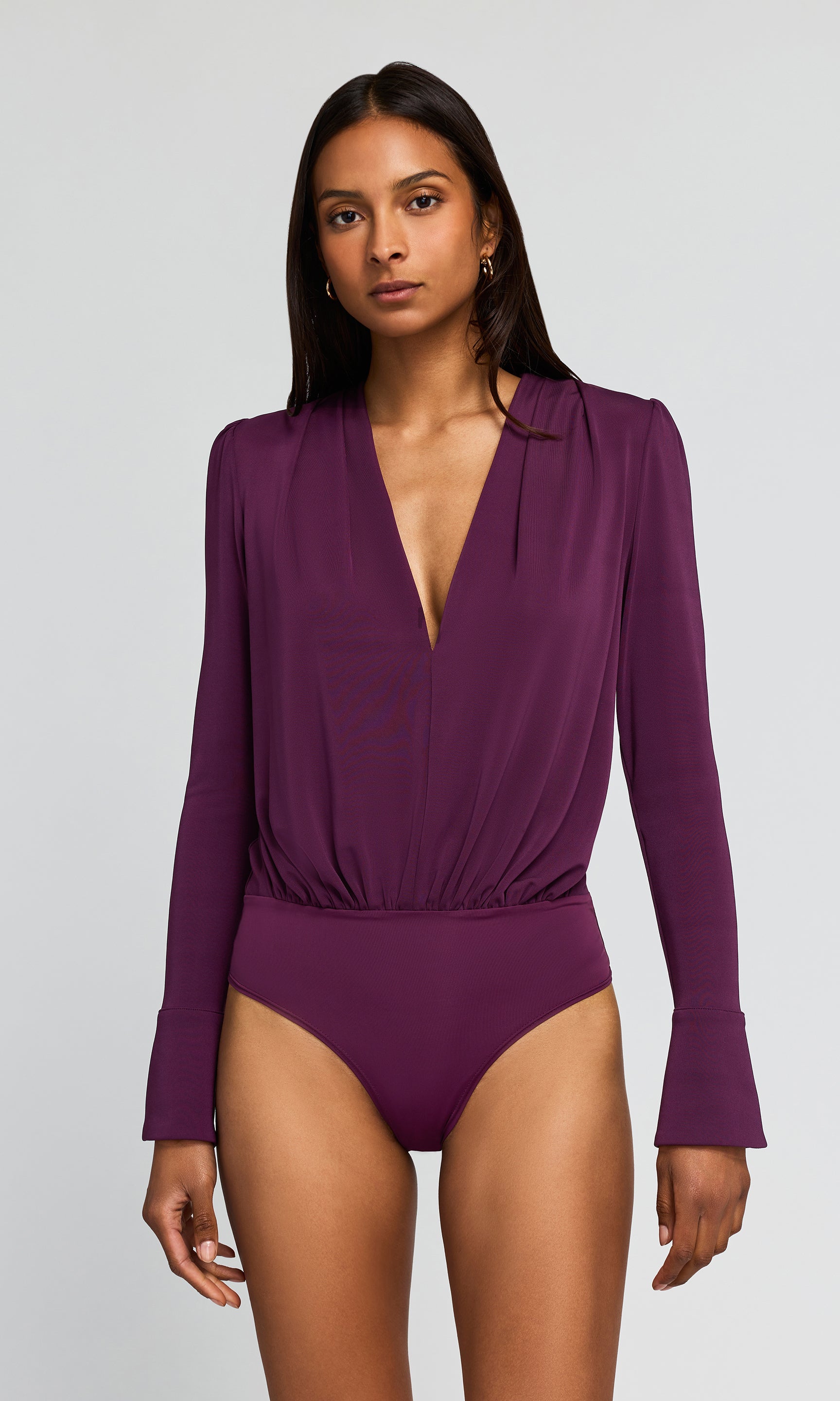 A woman modeling a plum purple long-sleeve one-piece bodysuit with a deep V-neckline and high-cut bottoms.
