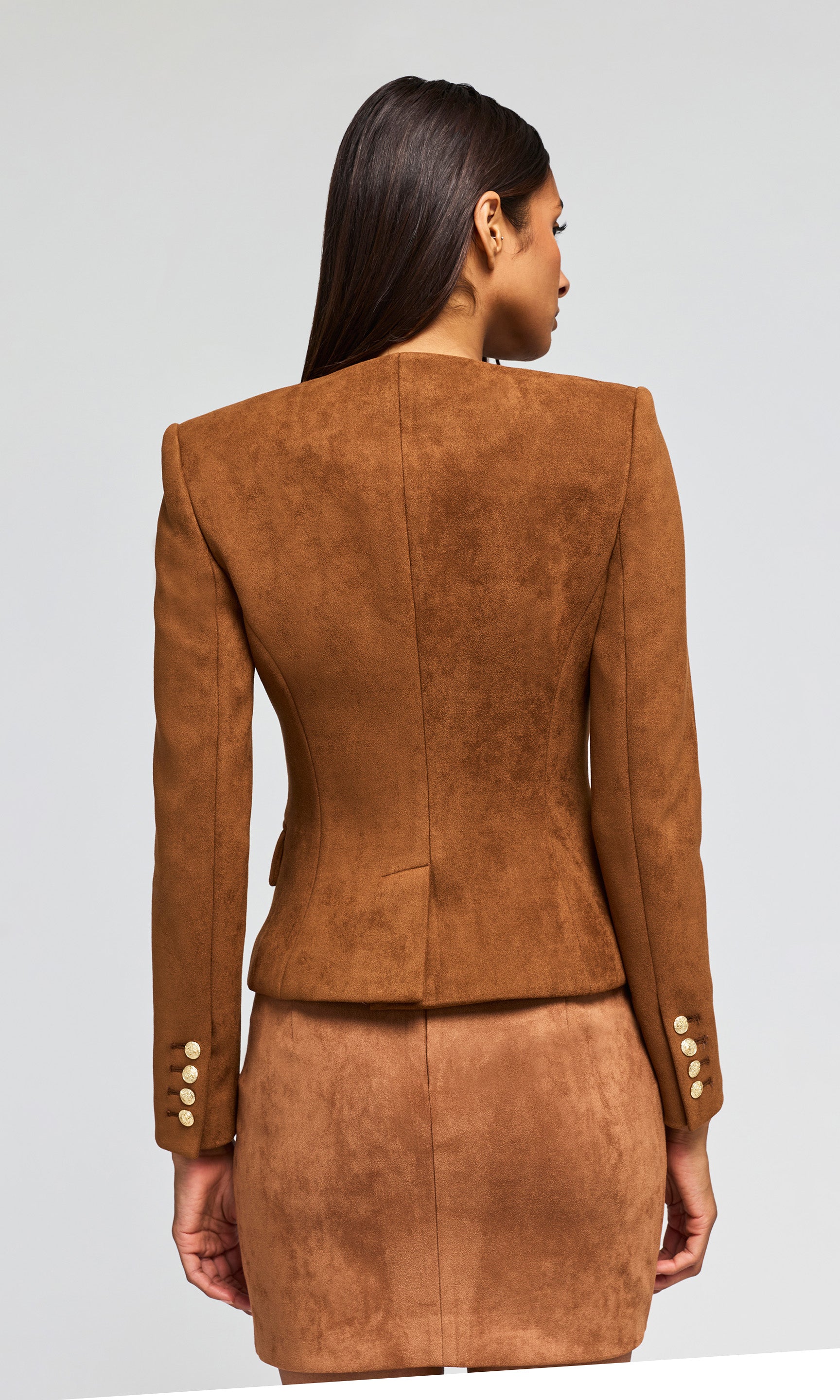 Back view of model wearing a brown suede-like two-piece suit consisting of a cropped jacket with gold buttons and a matching wrap skirt.