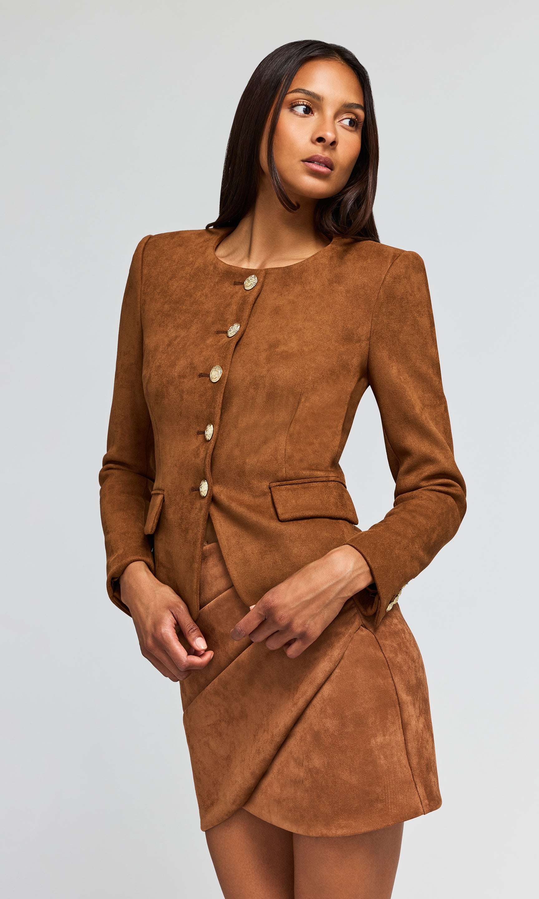 Side view of model wearing a brown suede-like two-piece suit consisting of a cropped jacket with gold buttons and a matching wrap skirt.