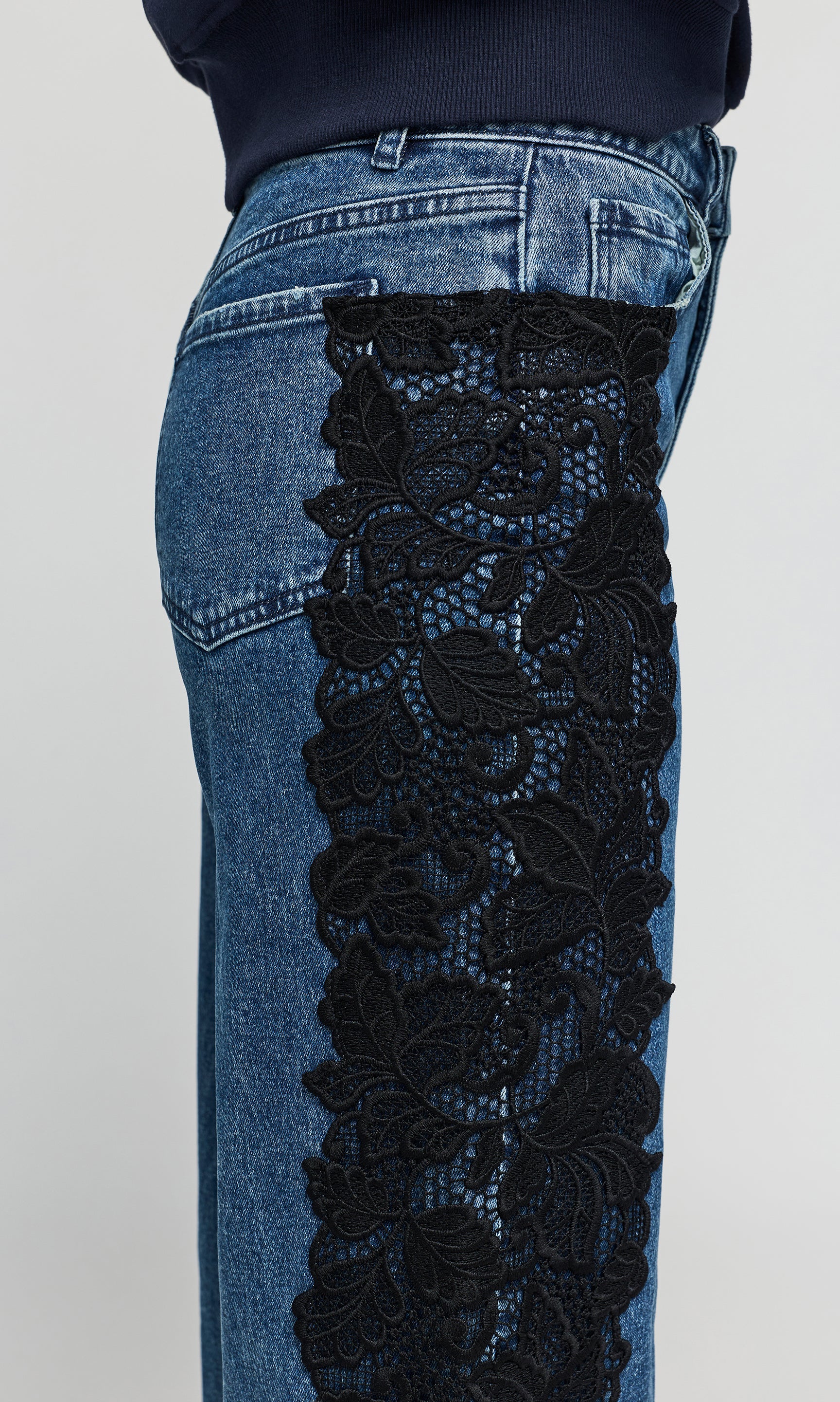 Close-up of blue denim jeans with a wide black floral lace panel attached down the side of the leg.