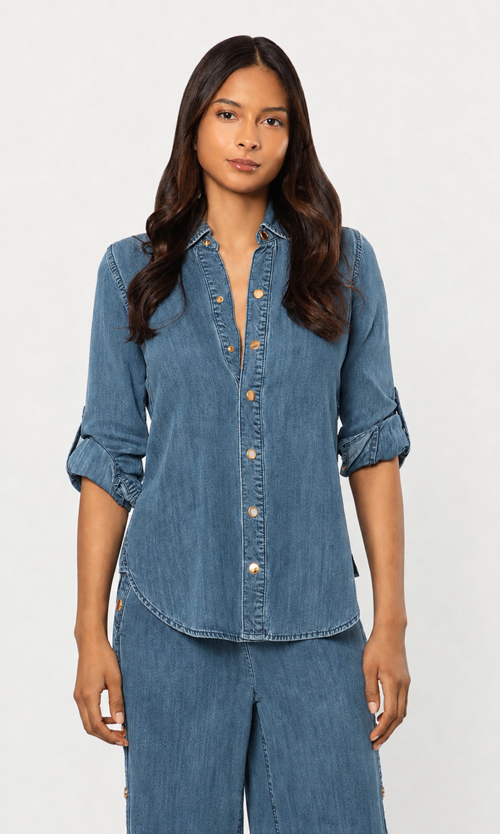 A woman modeling a blue denim button-up shirt with rolled sleeves and matching blue denim pants against a plain light gray studio background.