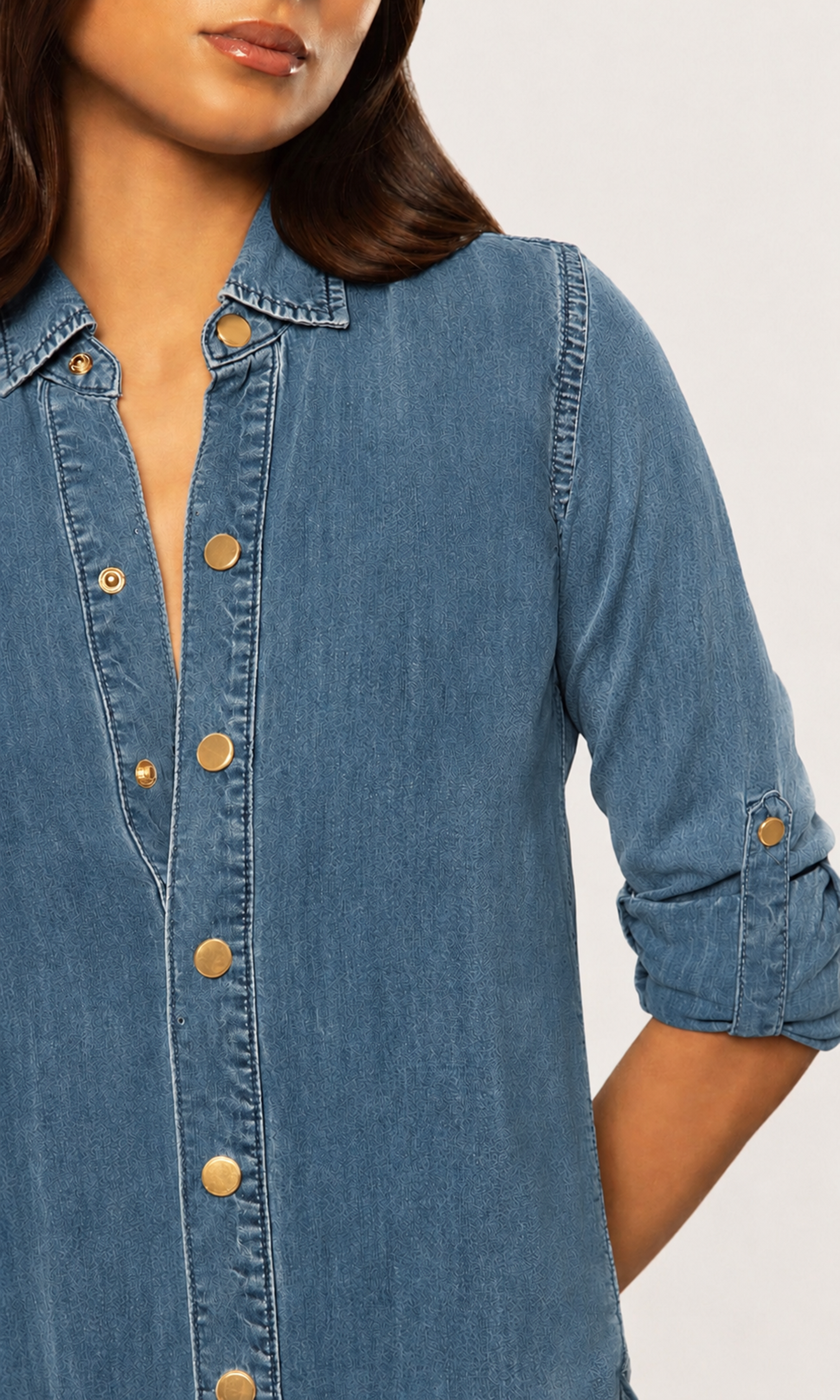 A close-up of a woman wearing a blue denim button-up shirt with rolled sleeves and gold-tone buttons, shown against a plain light background.
