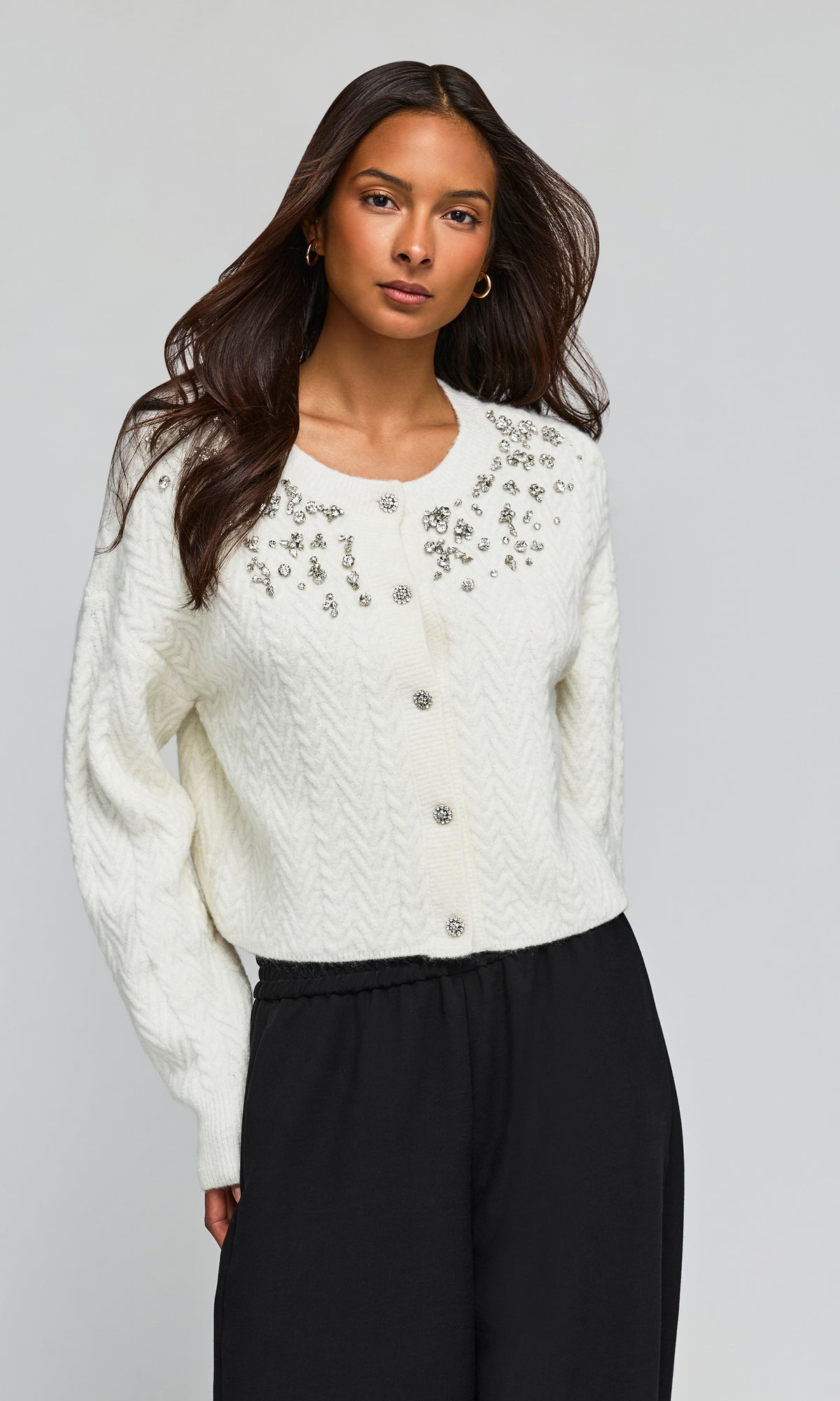 Side view of model wearing a cream cable-knit cropped cardigan with silver floral embroidery and button closures, paired with black pants.
