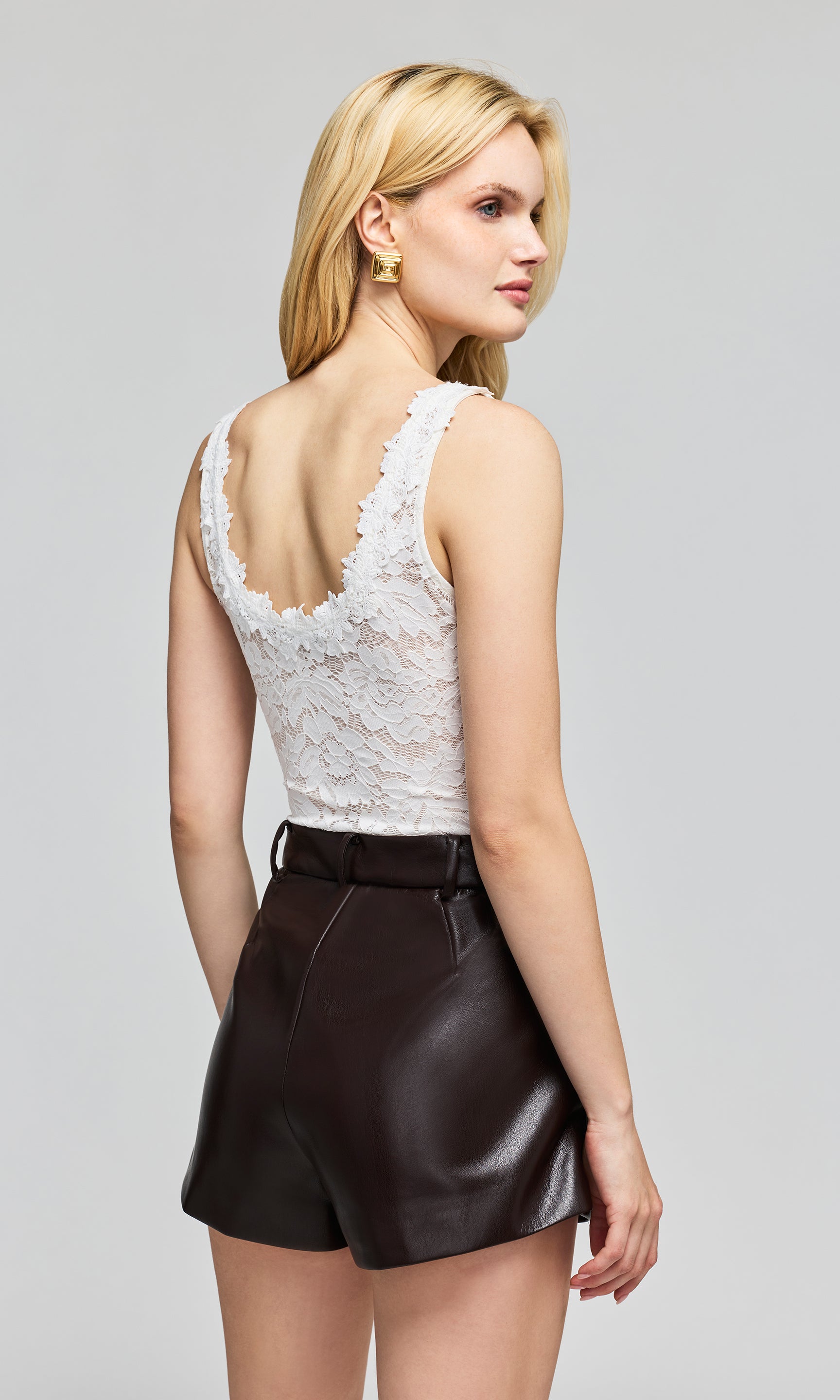 Back view of model wearing a white lace sleeveless bodysuit top and black faux-leather shorts, on a plain background.