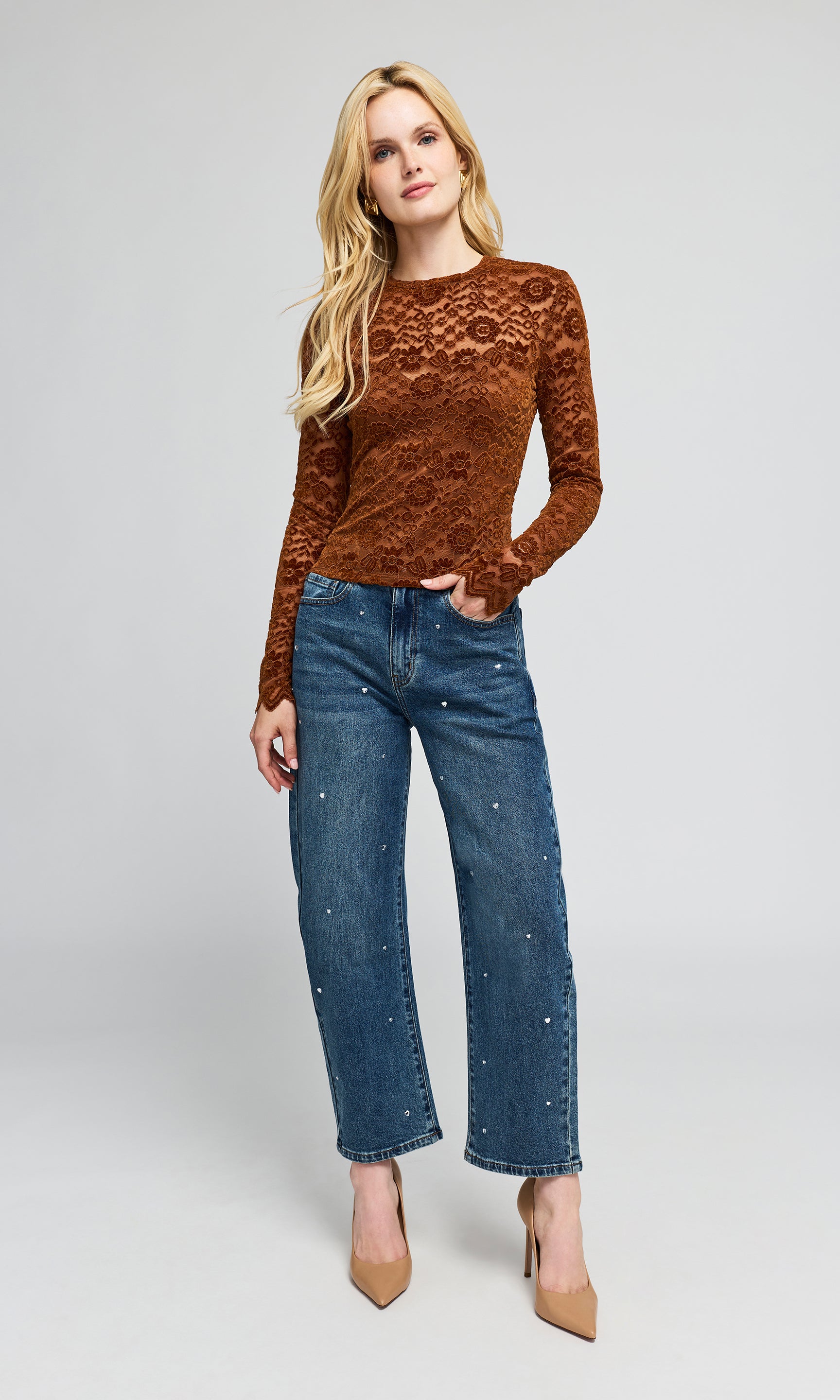 A blonde model wearing a rust-brown lace long-sleeve top, blue denim jeans with rhinestone embellishments.