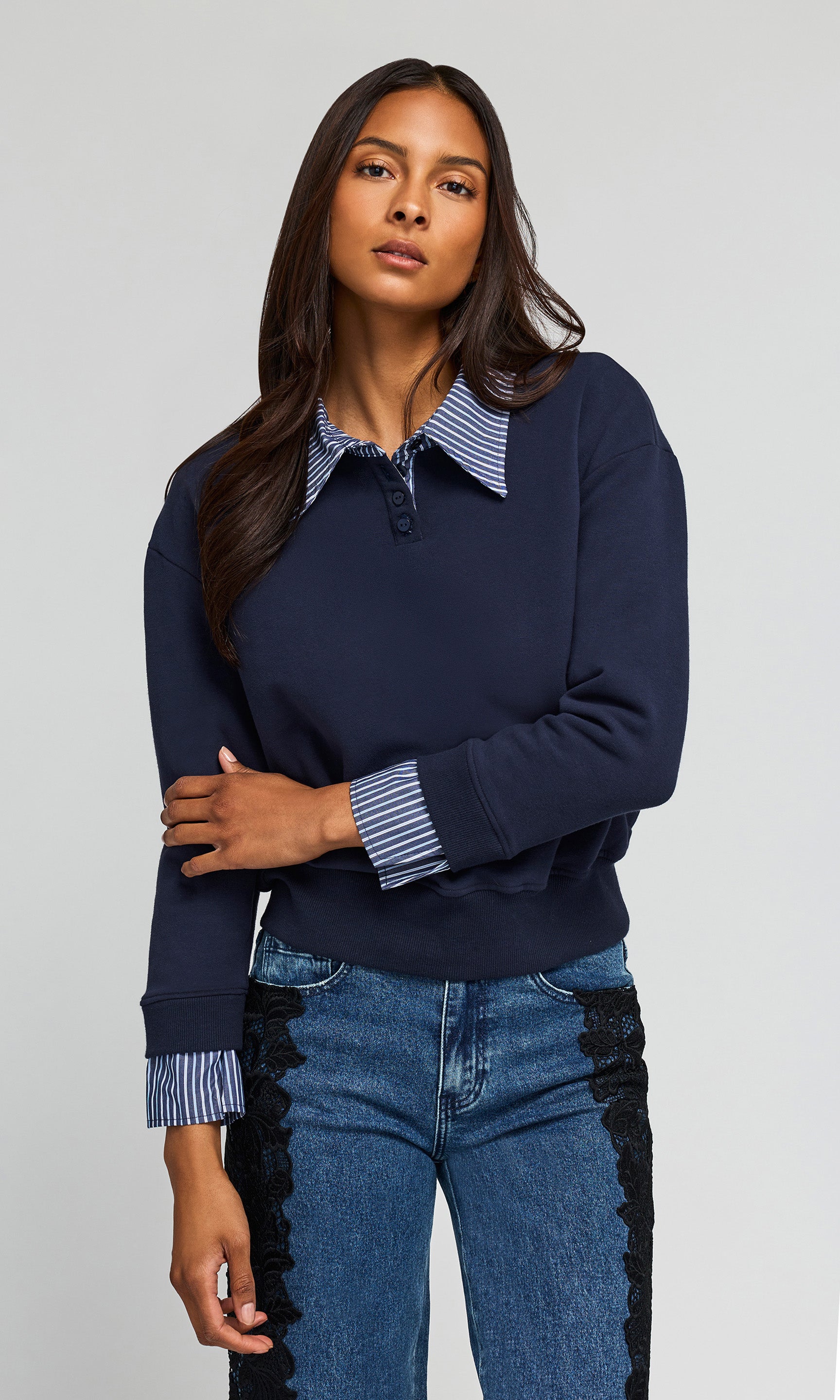 A woman modeling a navy blue long-sleeve knit top with a striped polo collar and striped button placket, paired with blue jeans against a light gray studio background.
