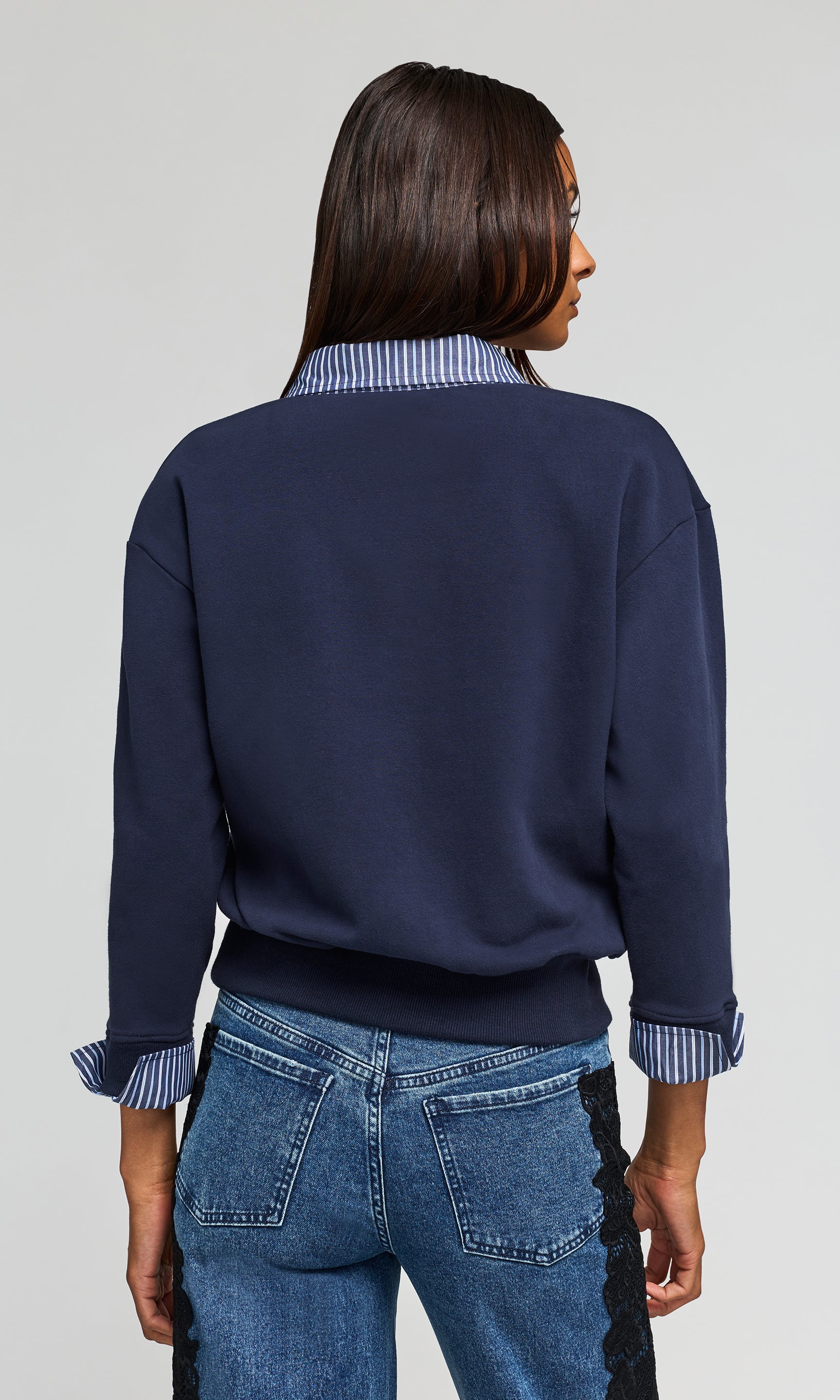 Back view of woman shown from the back wearing a navy blue sweatshirt with a blue and white striped collar, against a light gray studio background.