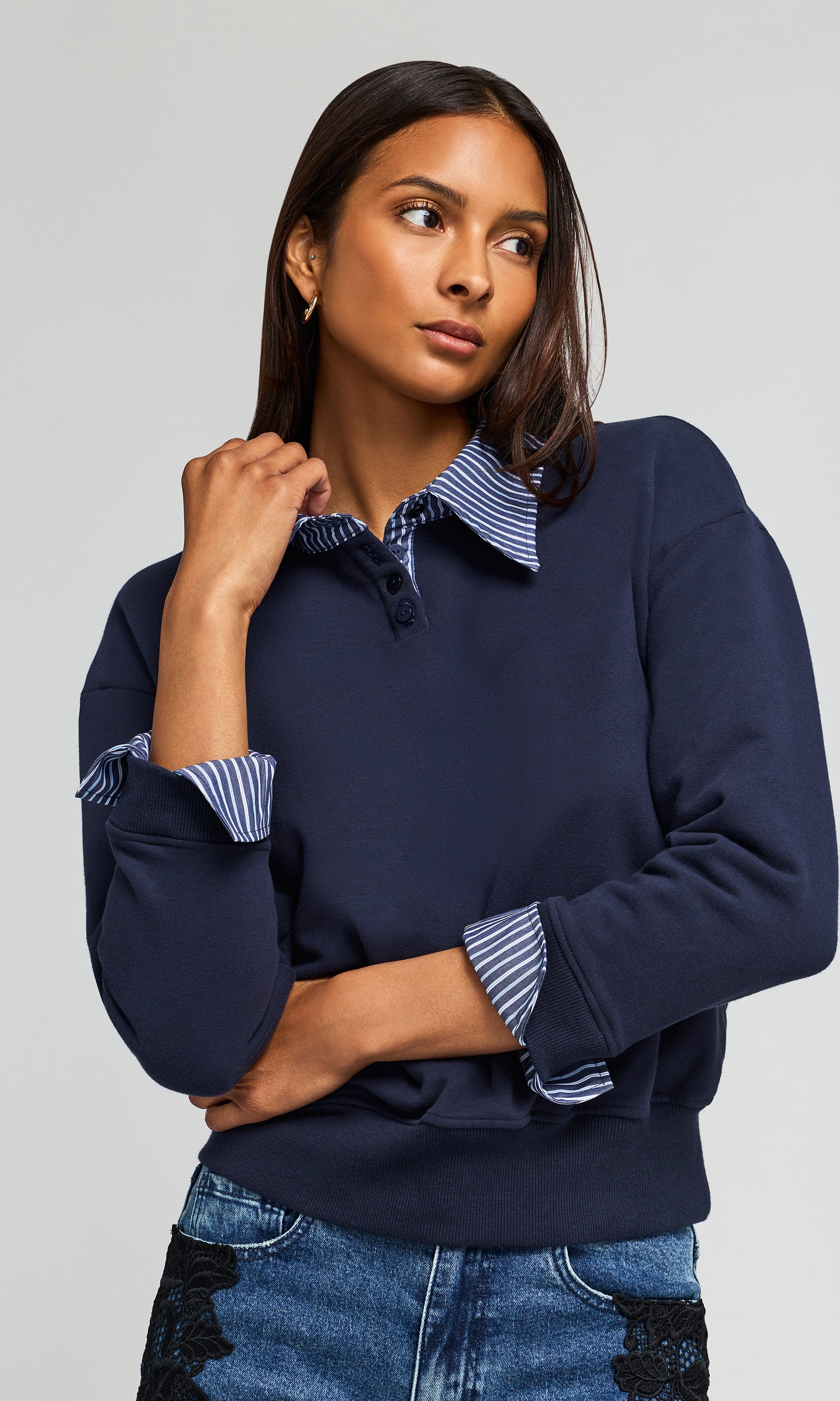 A woman wearing a navy blue long-sleeve top with striped button-up collar and striped rolled cuffs, paired with blue jeans, against a light gray studio background.