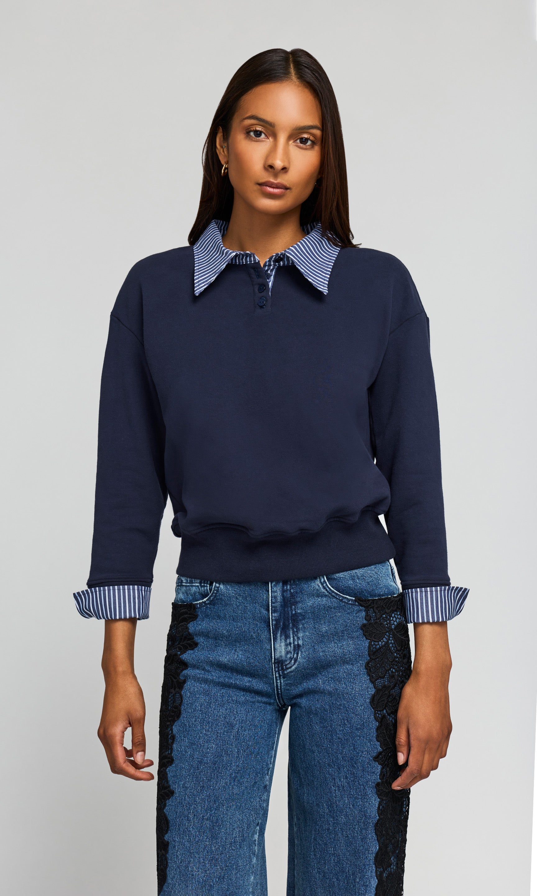 Model wearing a blue long-sleeve knit top with a contrasting blue and white striped polo-style collar and striped turned-back cuffs, paired with blue jeans on a plain background.