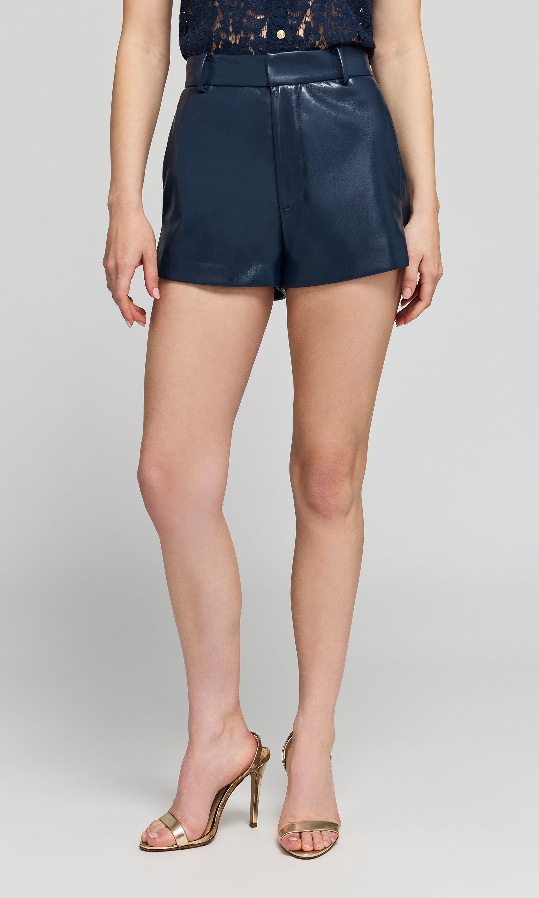 A model wearing navy blue faux-leather shorts, paired with metallic gold high-heeled sandals on a plain background.