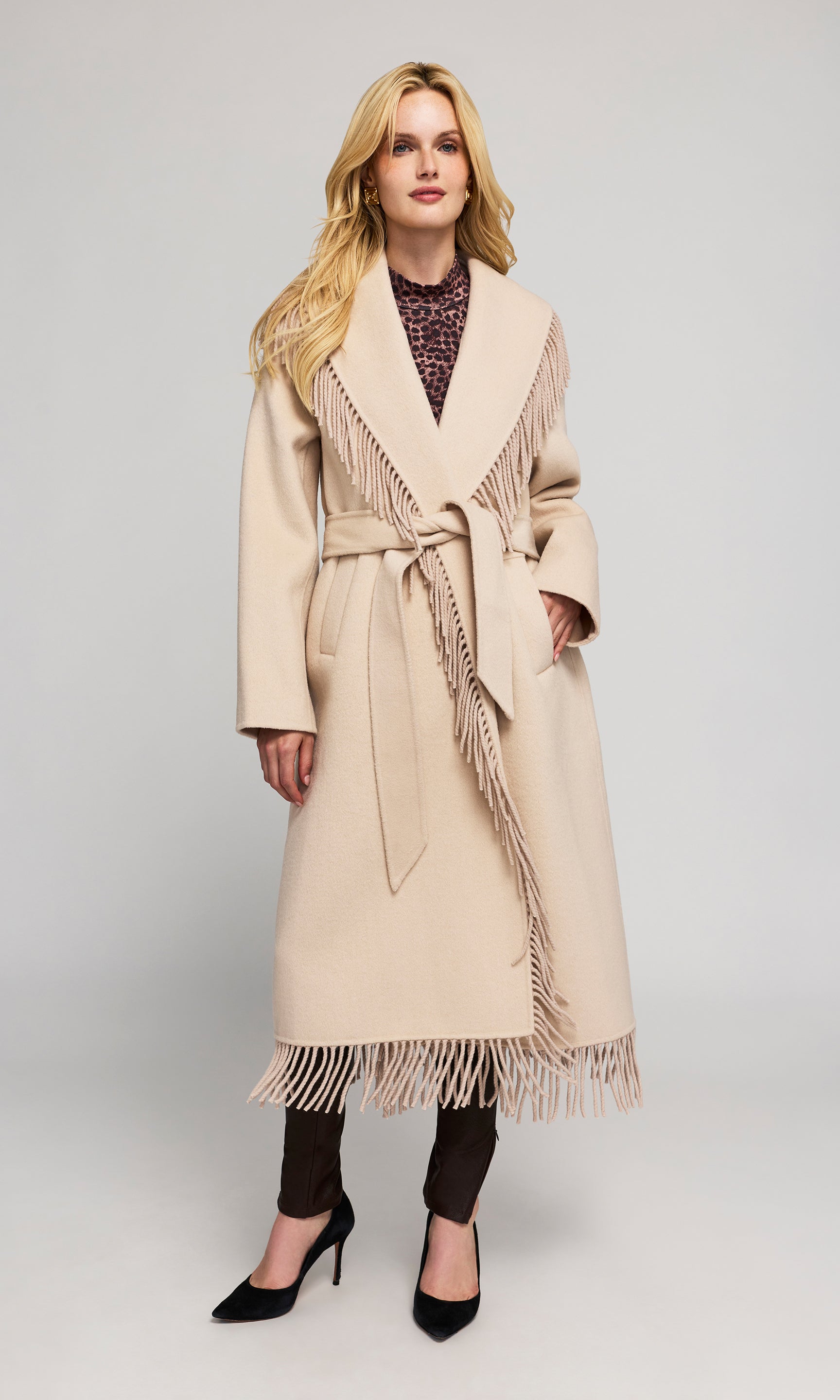 Model wearing a long beige coat with fringe trim and a tie belt, paired with black tights and black high heels.