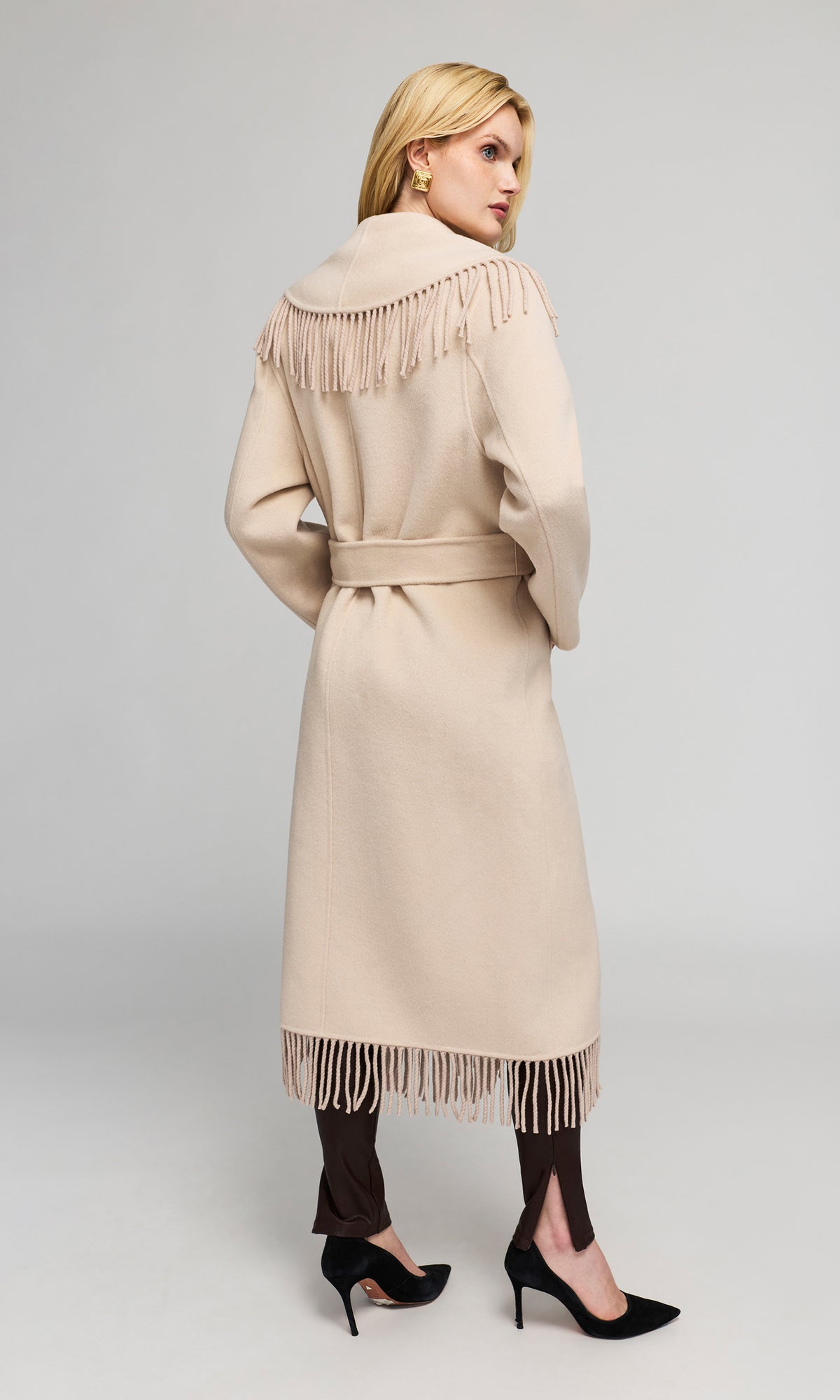 Back view of model wearing a long beige coat with fringe detailing on the shoulders and hem, paired with dark brown tights and black heels.
