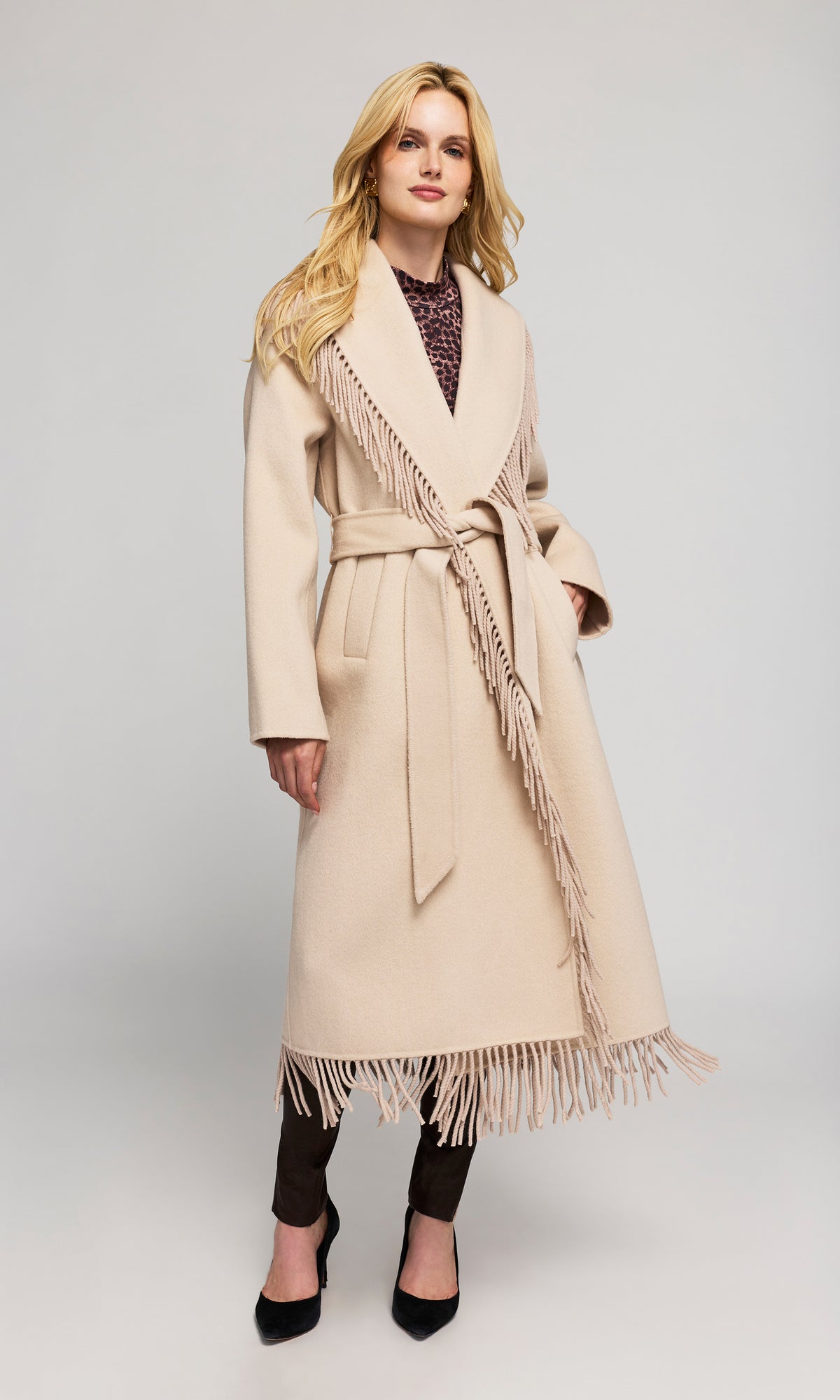 Side view of model wearing a long beige coat with fringe trim and a matching tie belt.