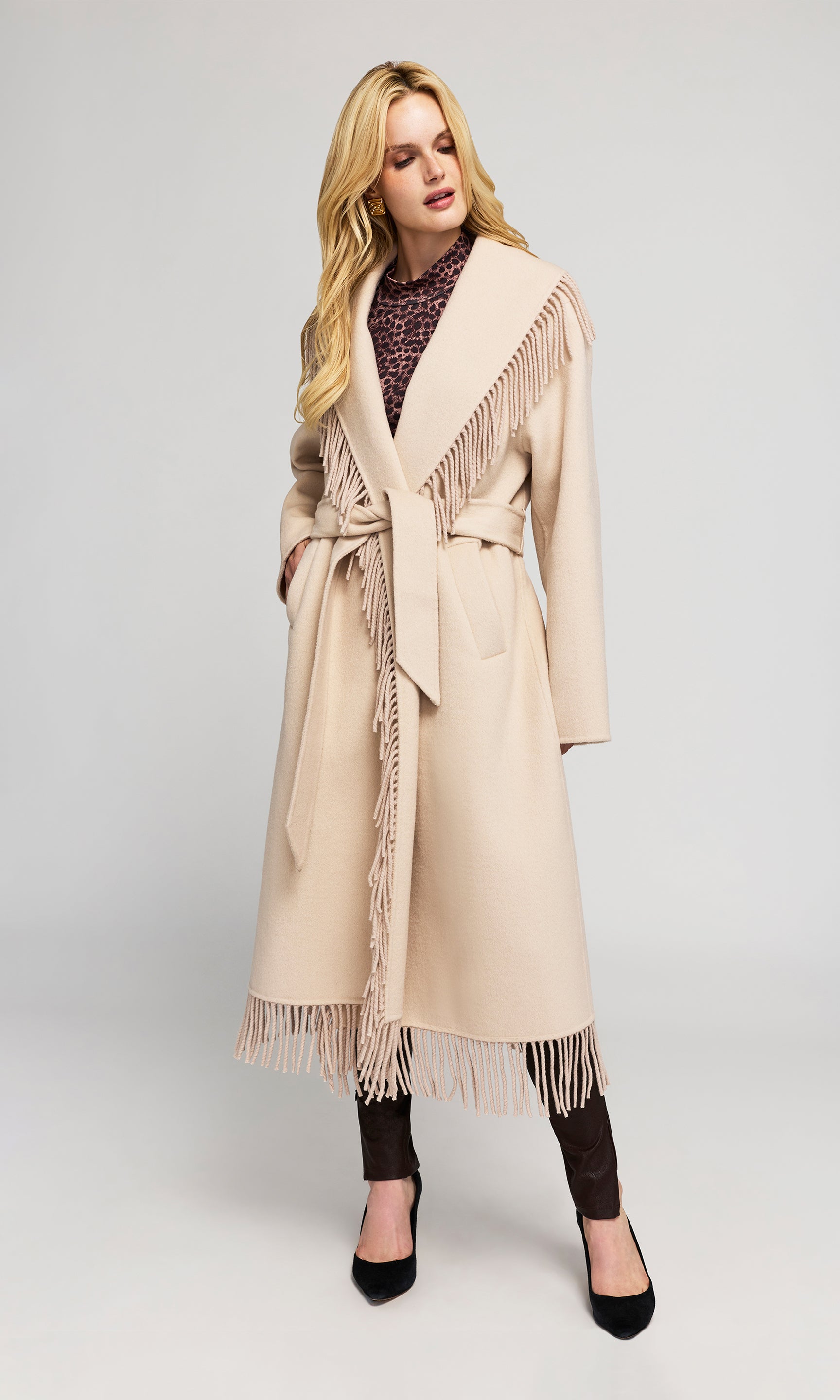 Model wears a long beige wrap coat with fringe trim, a matching waist tie belt, and large shawl-style lapels, styled over a dark patterned top, dark leggings, and black heels.