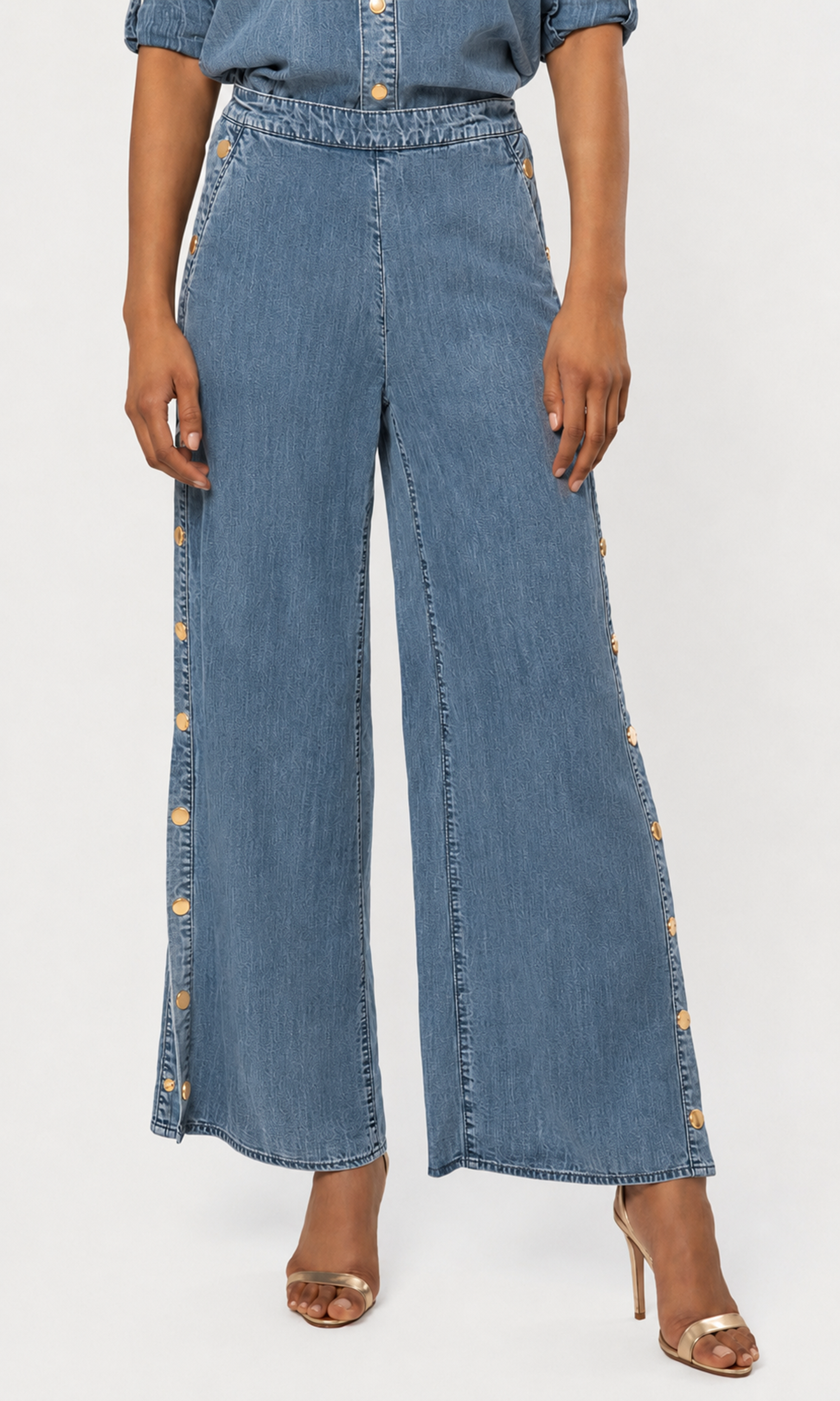 Model wearing high-waisted, wide-leg denim pants with decorative gold button details running down the sides, paired with a blue denim top.