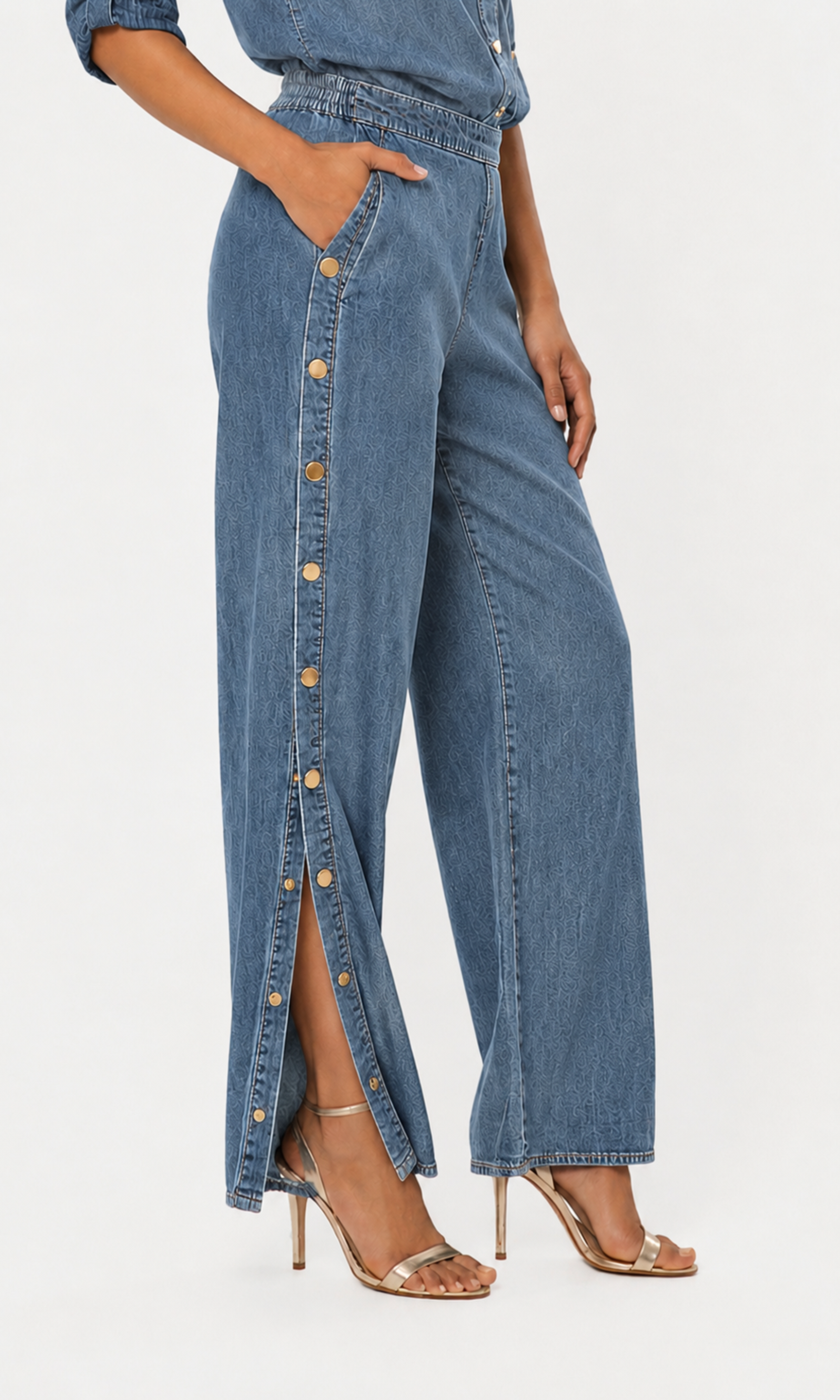 Side view of model wearing high-waisted, wide-leg blue denim pants with a vertical row of gold-tone buttons down side and a high side slit.