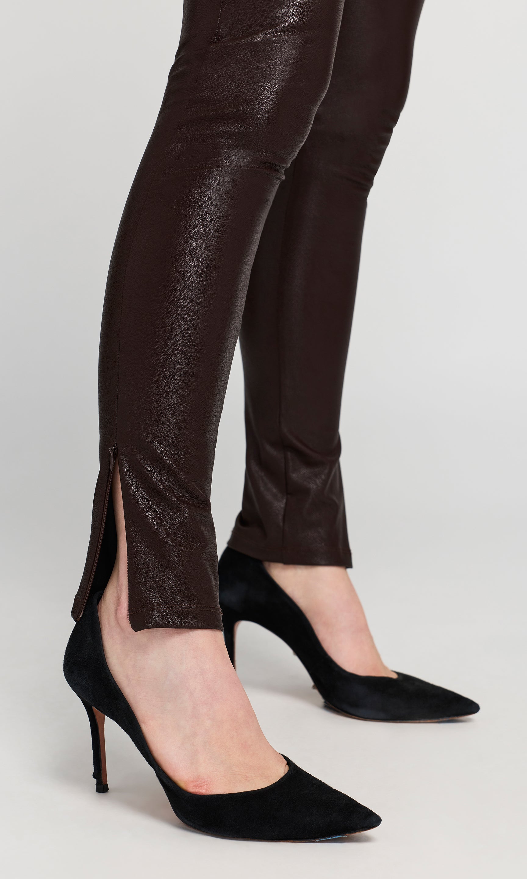 A close-up of a person's legs wearing dark brown faux-leather leggings with ankle slits.