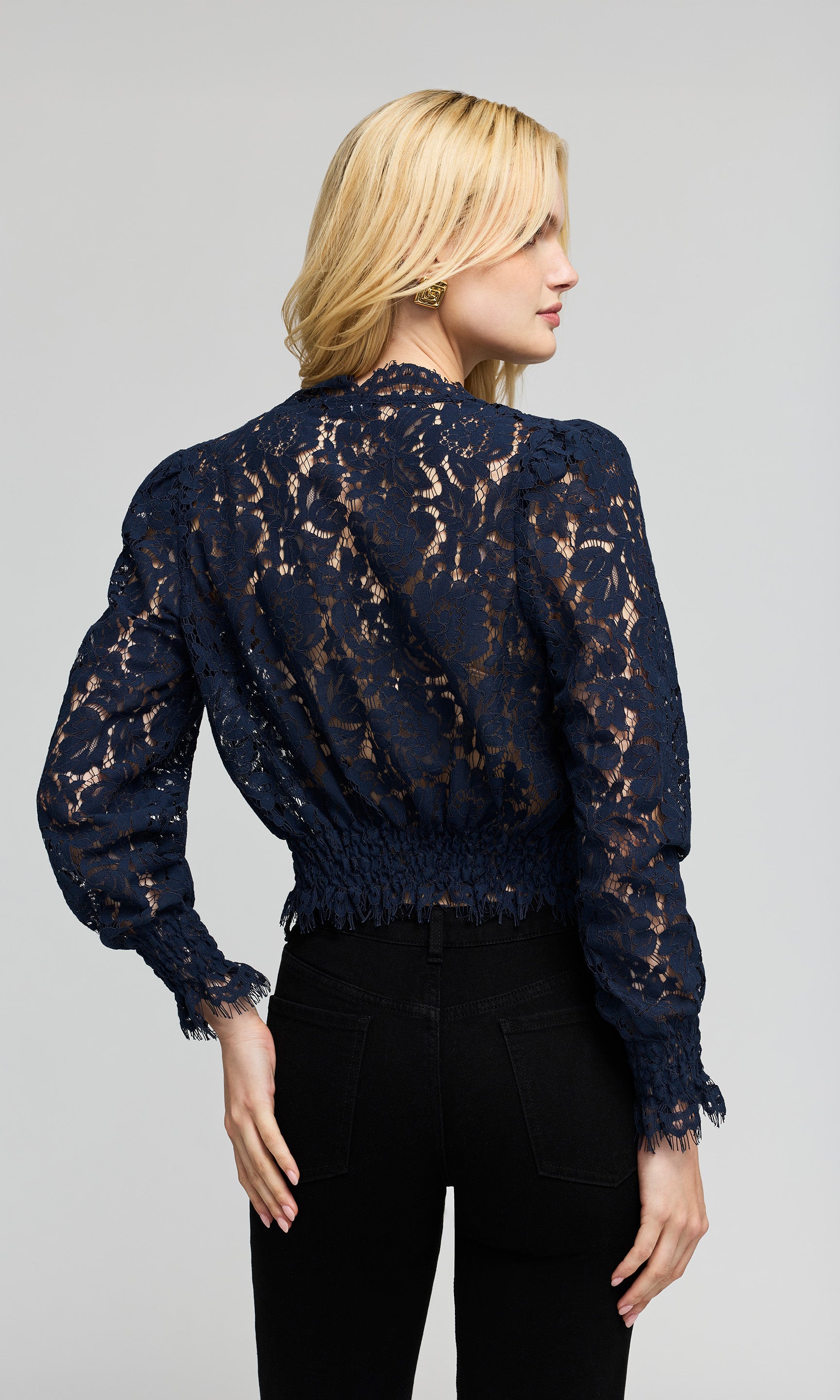 Model shown from the back wearing a navy blue lace long-sleeve blouse with a smocked waist and black pants.