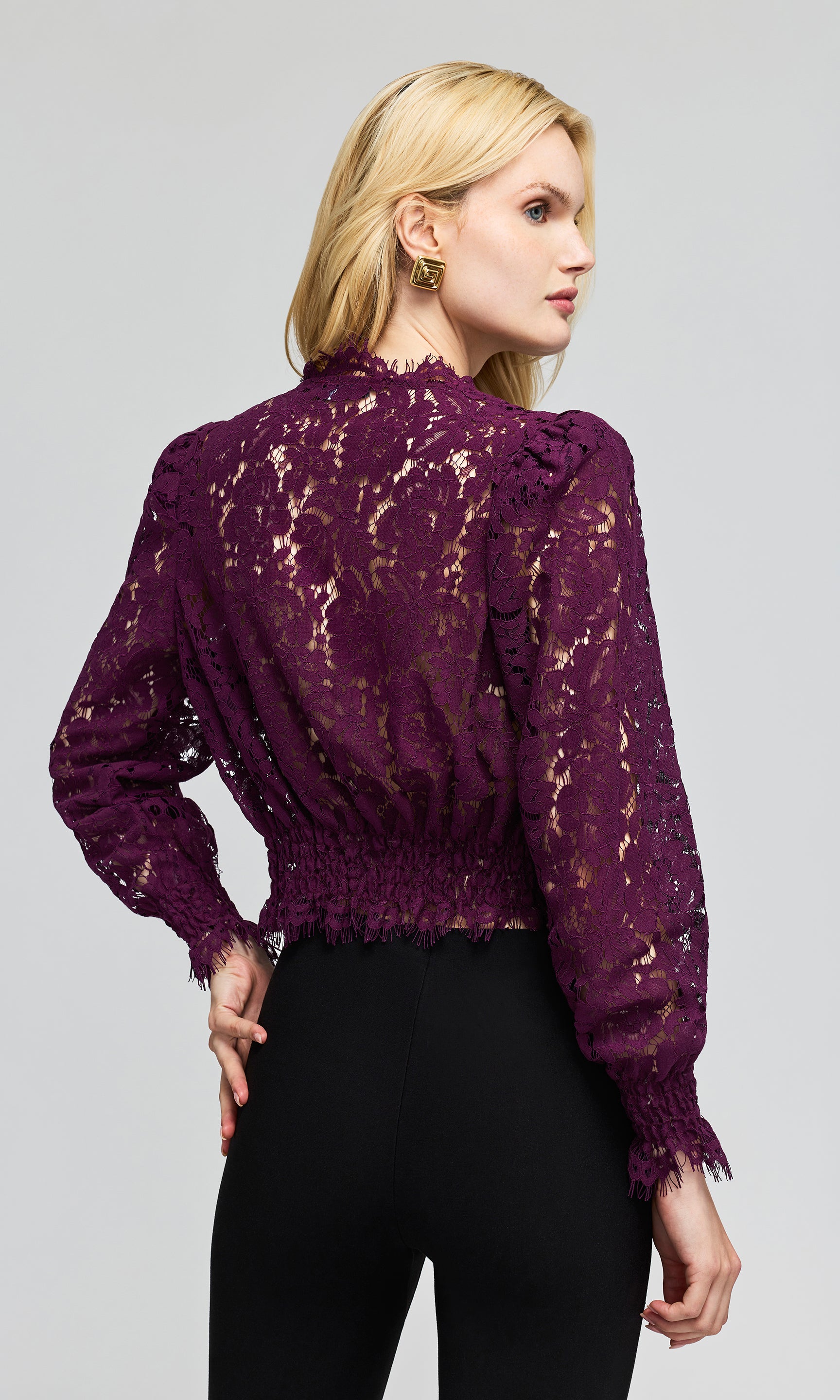 Back view of model wearing a deep purple lace long-sleeve blouse with a deep V-neck and black pants, on a plain background.